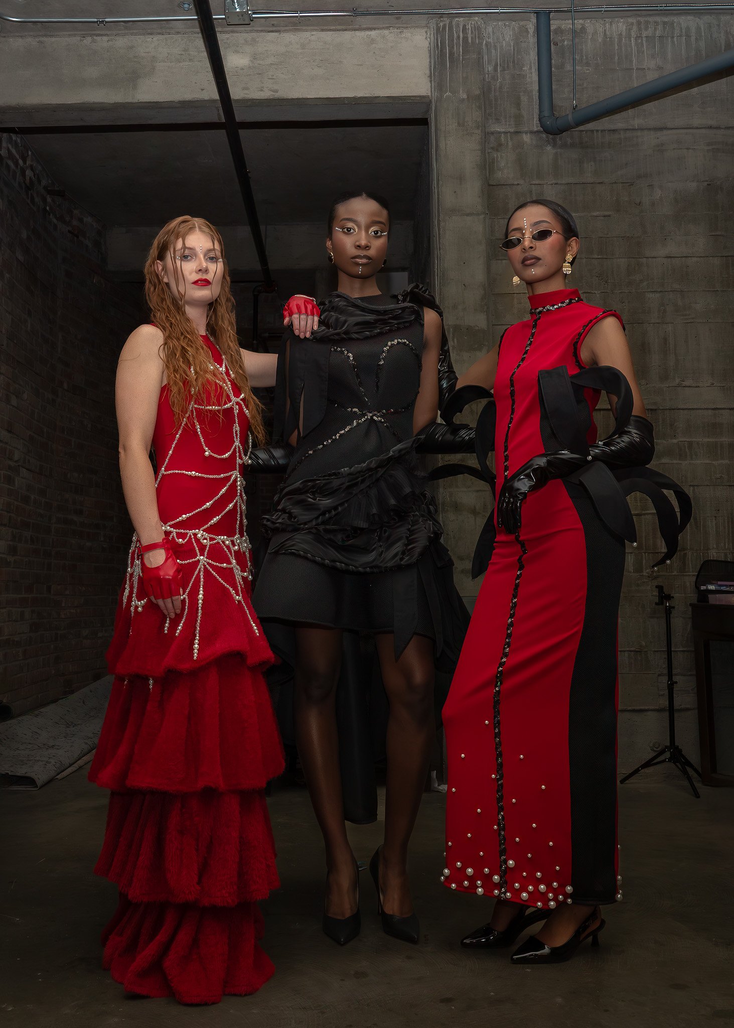 Three women standing together in a concrete setting. The woman on the left has long red hair and is wearing a red gown with silver chains. The woman in the middle has dark skin and black hair, dressed in a black dress with sheer fabric and black glov