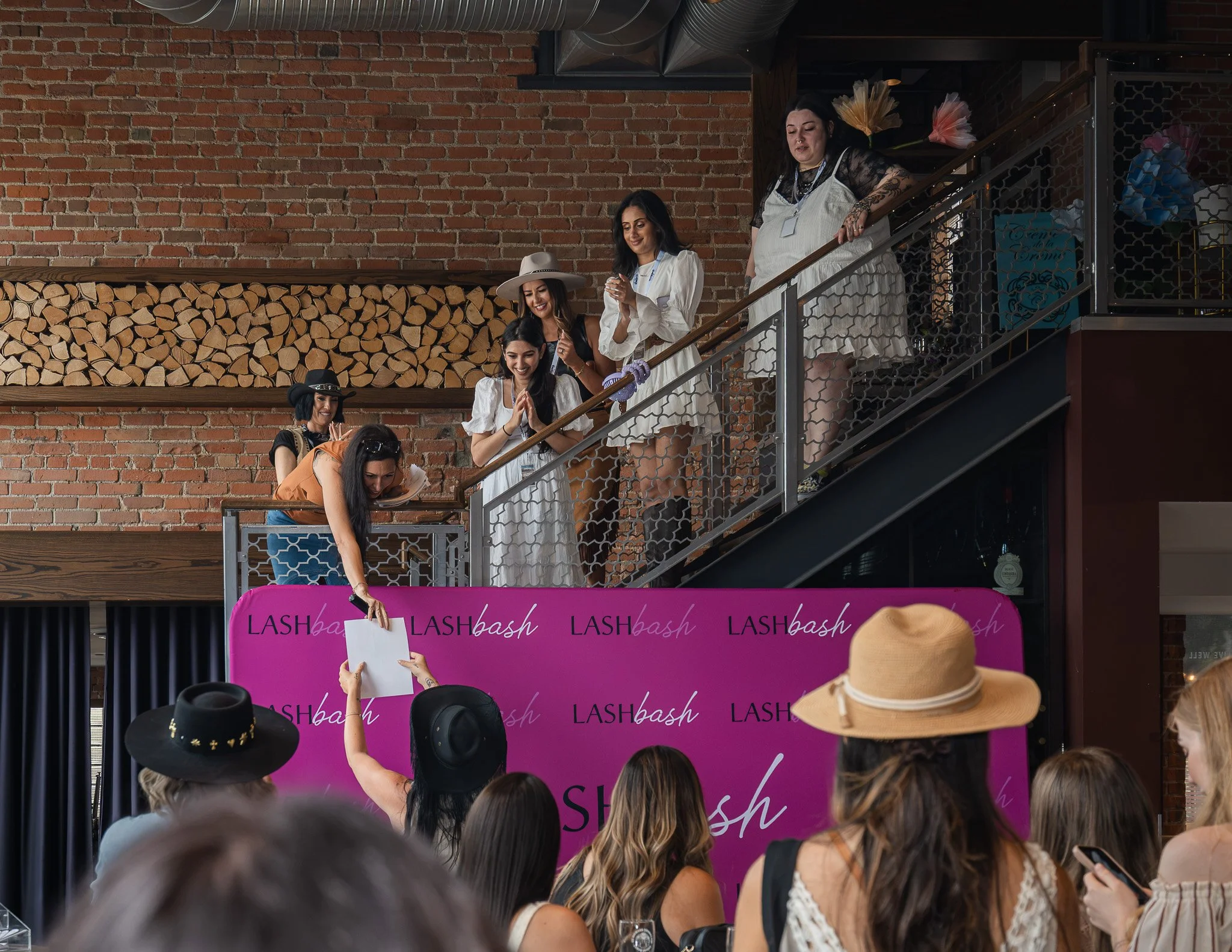 Group of women standing on a staircase, some with hats, at a beauty event with a purple backdrop that reads 'LASH bash,' in front of an audience.