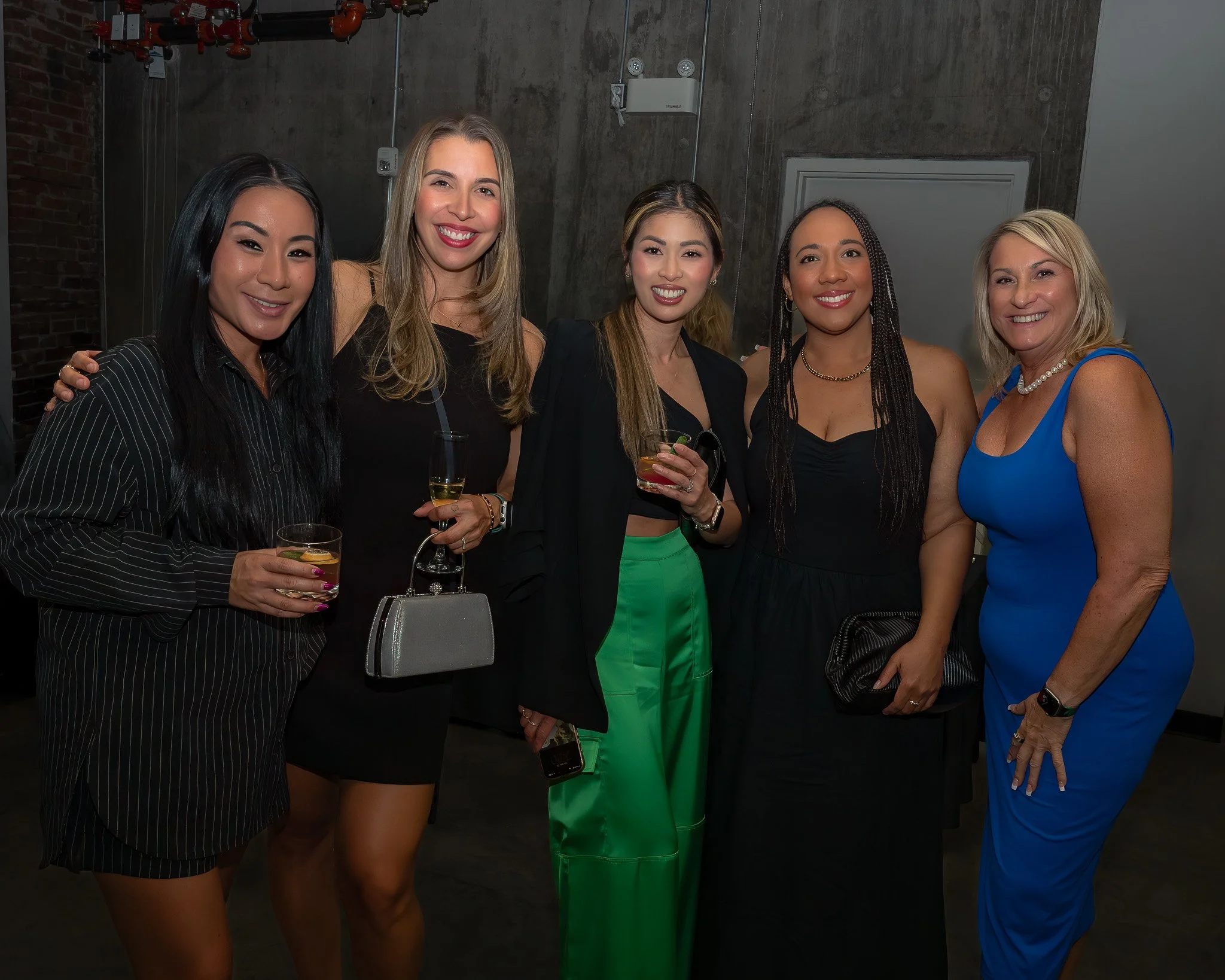 A group of five women posing for a photo at a social event, holding drinks and smiling. They are dressed in stylish attire, standing in front of an industrial-style concrete wall with red piping overhead.