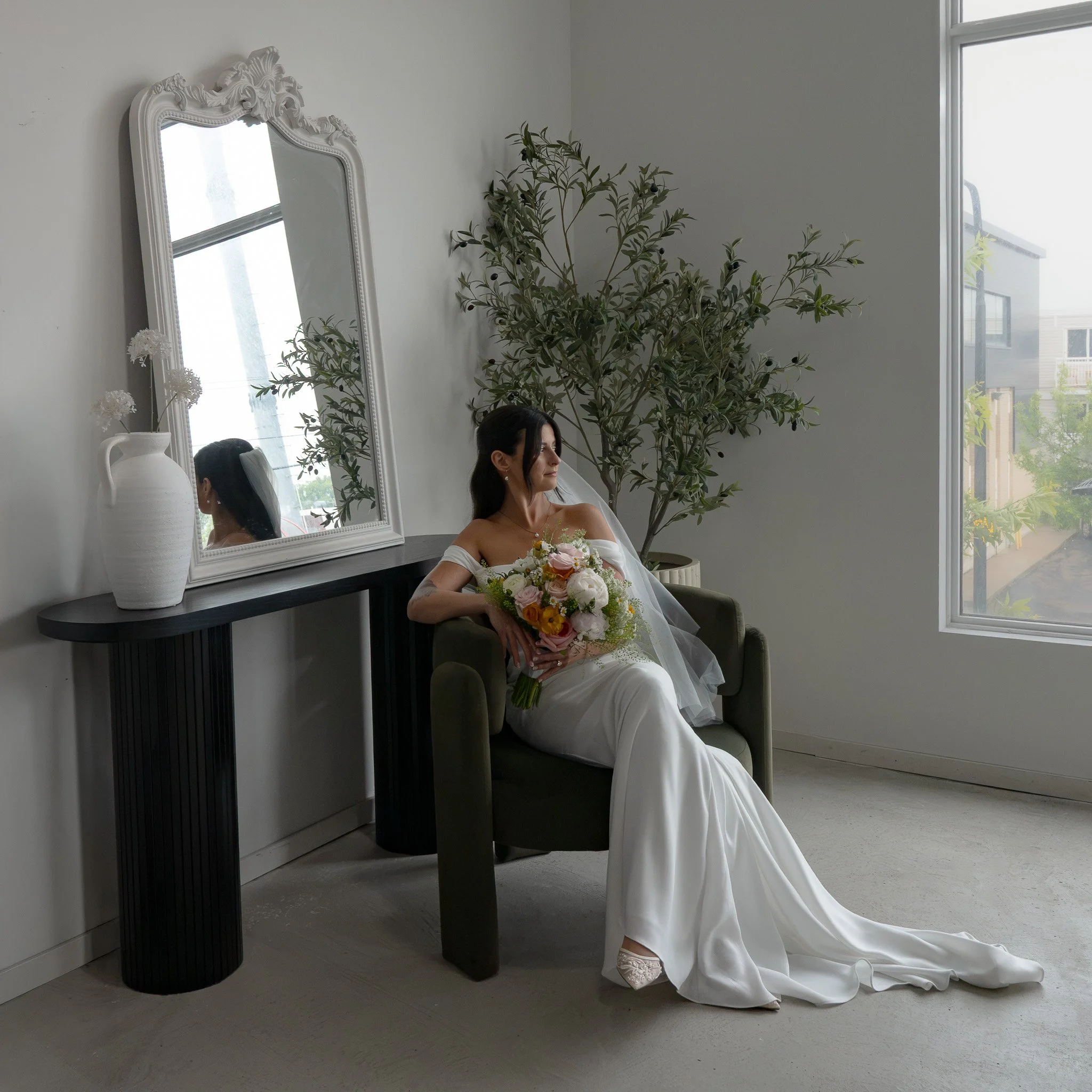 A woman in a wedding dress sitting on a green chair in a room with white walls, a large mirror, a tall white vase with flowers, and a potted plant near the window.