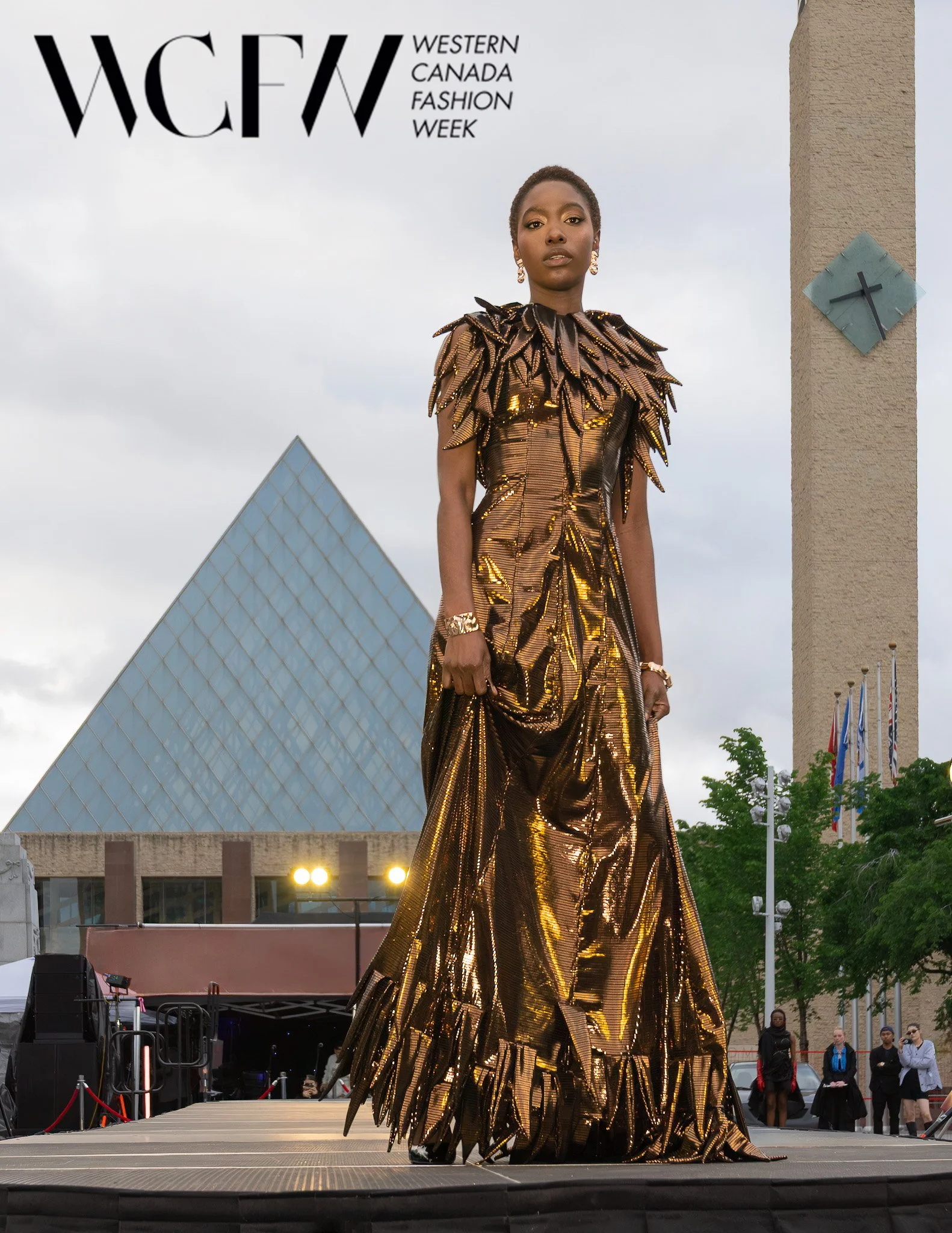 A woman in a gold metallic dress with leaf-shaped embellishments standing on a stage at Western Canada Fashion Week, with a modern building and cloudy sky in the background.