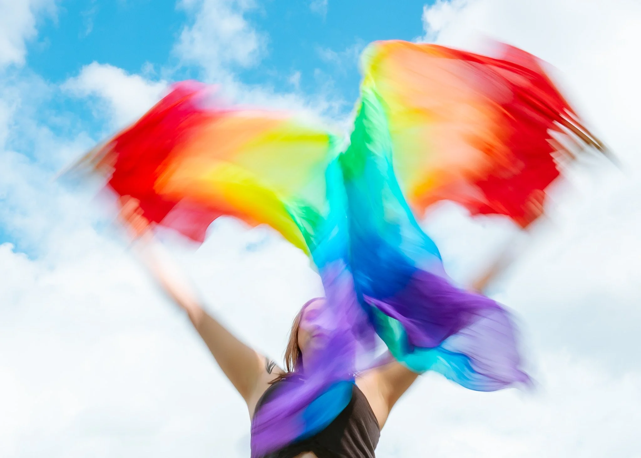 Person holding colorful rainbow flags up in the air outdoors against a cloudy sky.