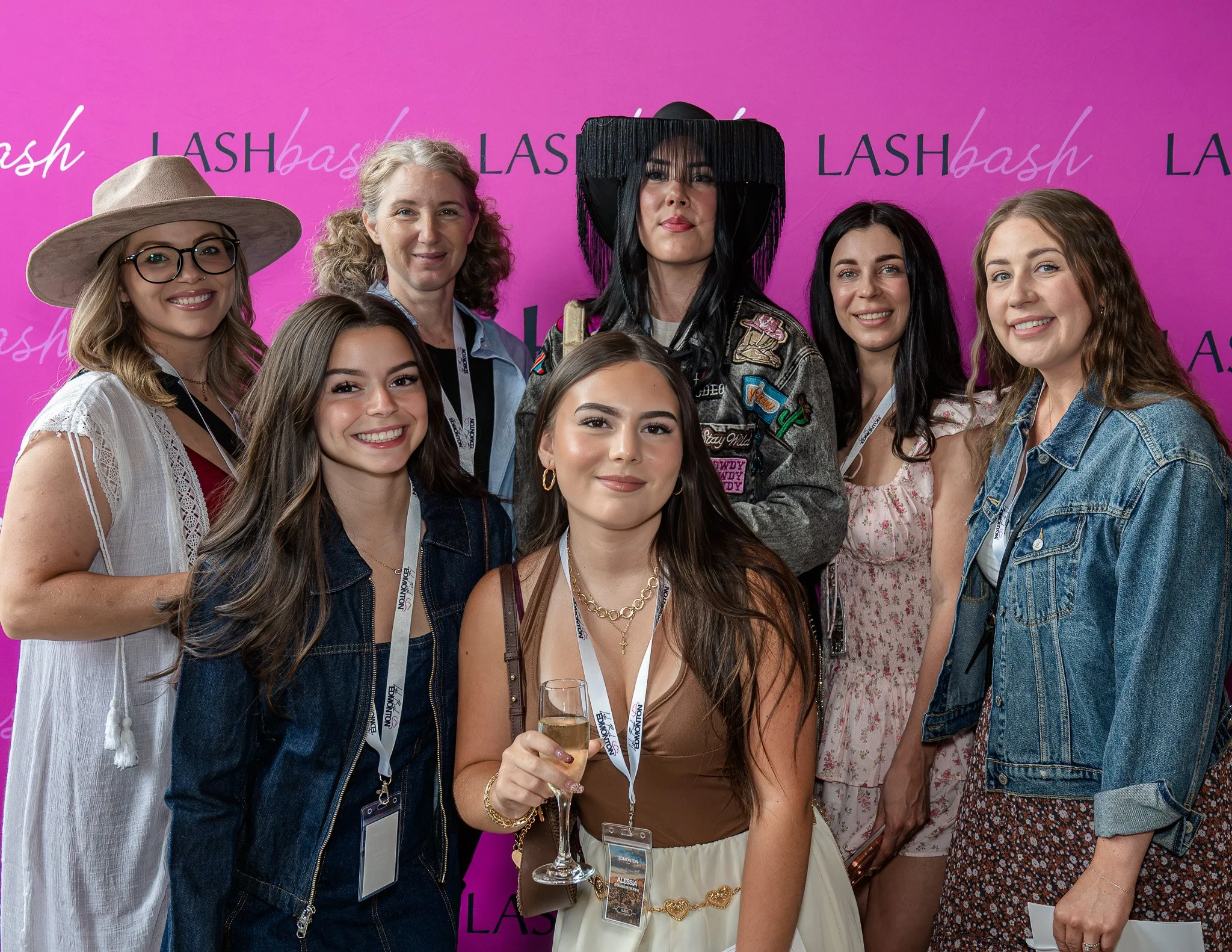 Group of women and young women at an event, standing in front of a purple backdrop with the word 'LASHbash' repeated. One woman holds a glass of champagne.