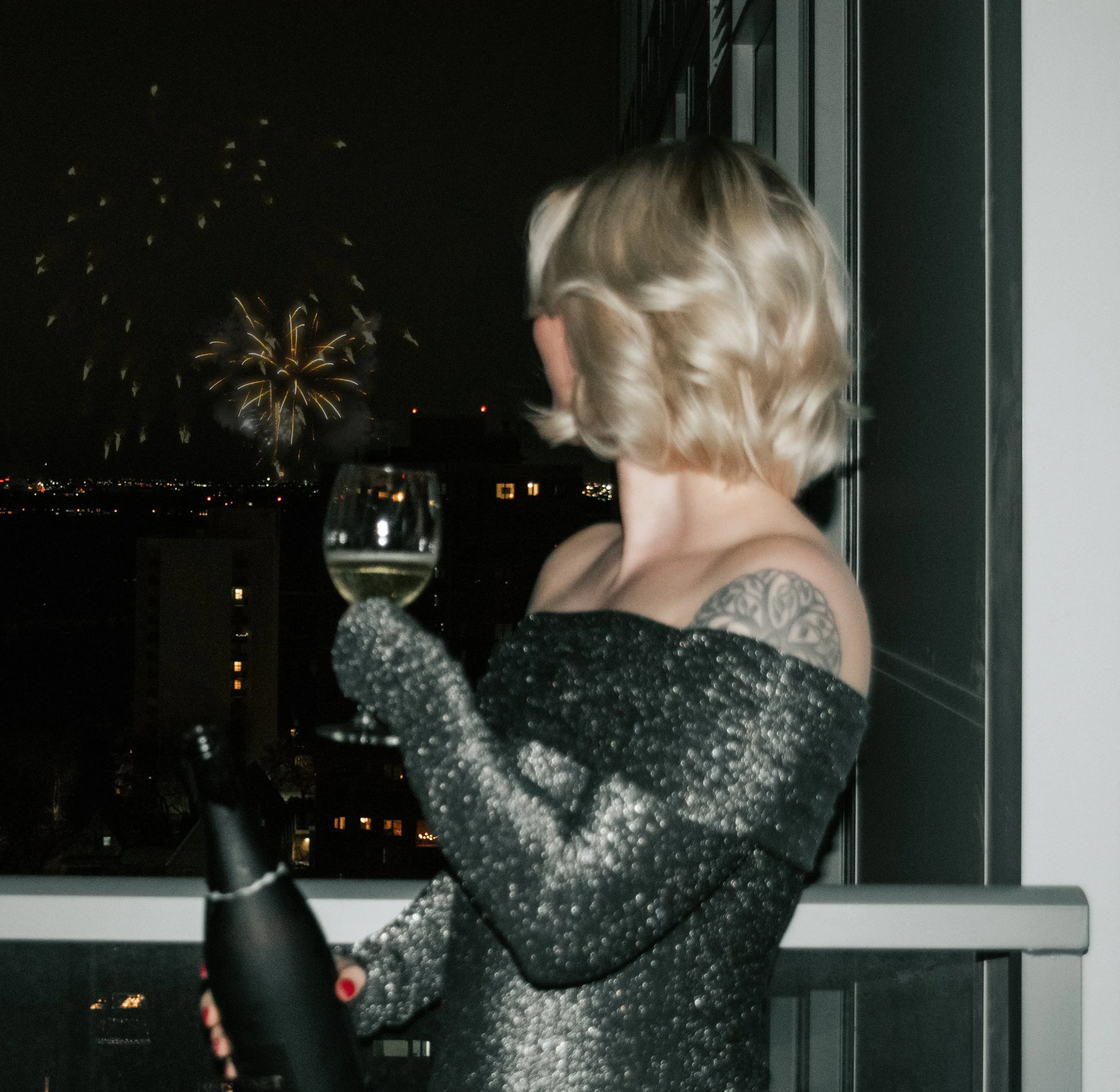 A woman with short blonde hair, wearing an off-the-shoulder black and gray sparkly dress, holds a glass of white wine while looking at fireworks outside a window at night.