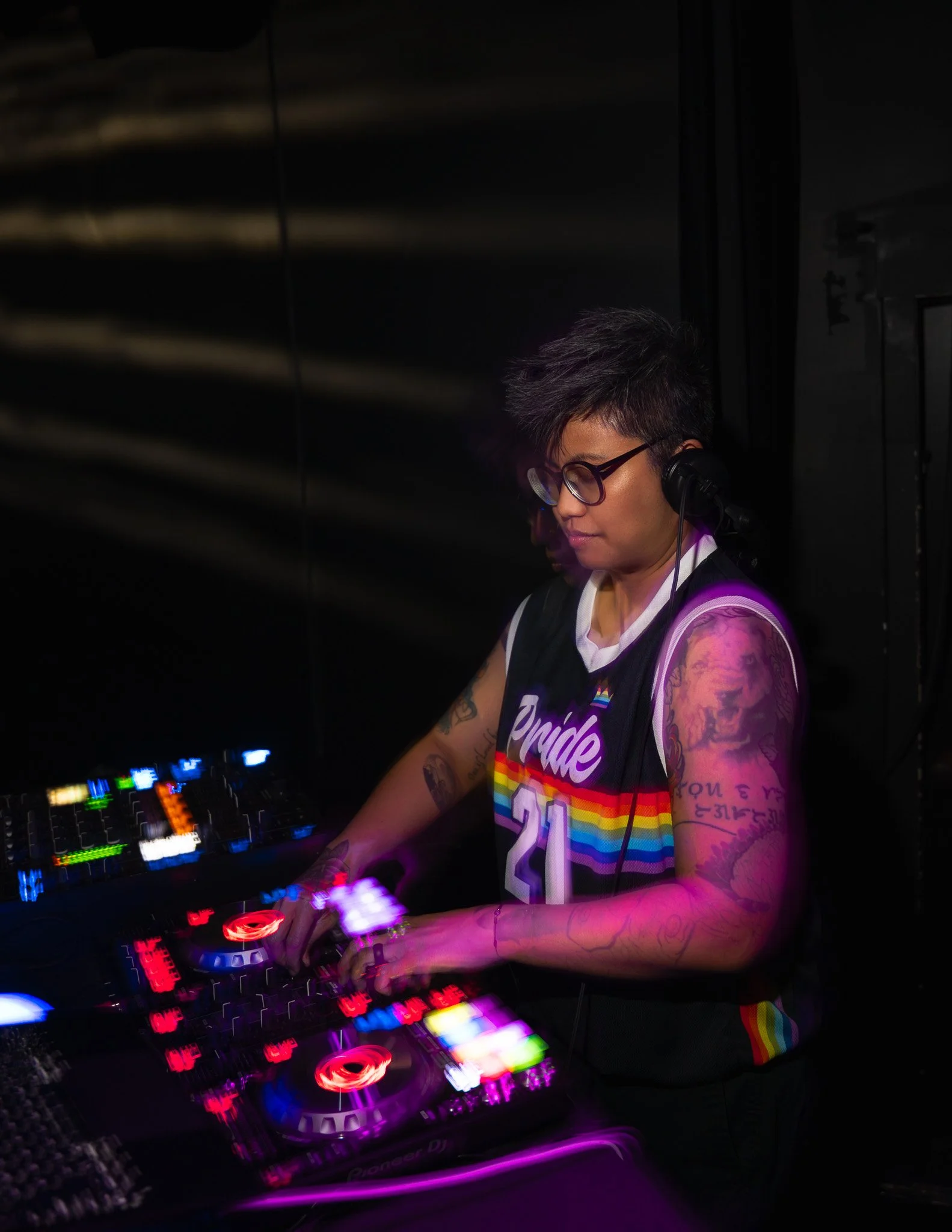 A DJ with short dark hair and glasses, wearing a rainbow-colored pride shirt, is mixing music at a DJ controller with colorful LED lights, in a dark setting.