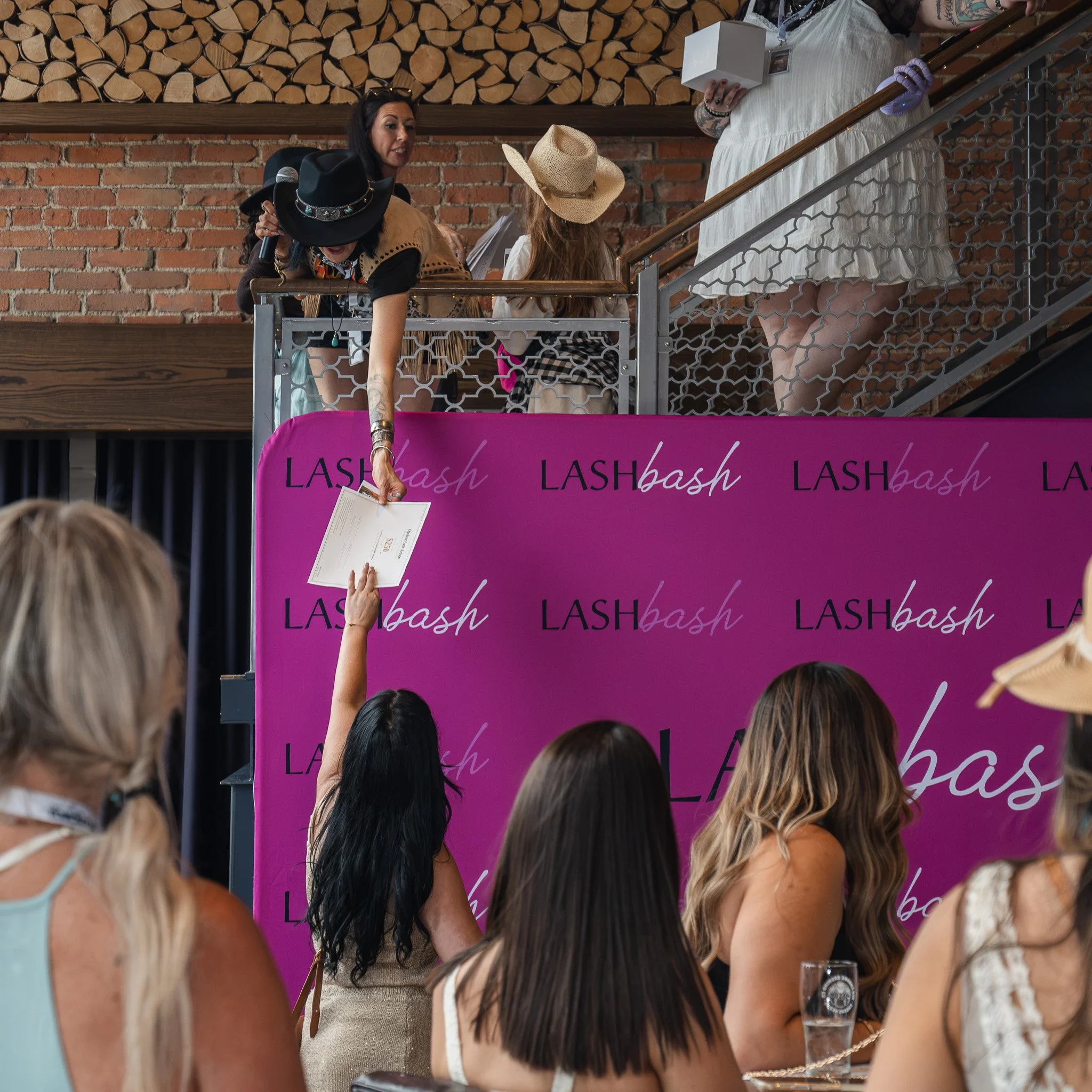 A group of women at a Lash Bash event, standing behind a pink booth with the event name on it. Some women are wearing hats, and one woman is holding a certificate or voucher. The background features a brick wall with logs stacked on the upper section