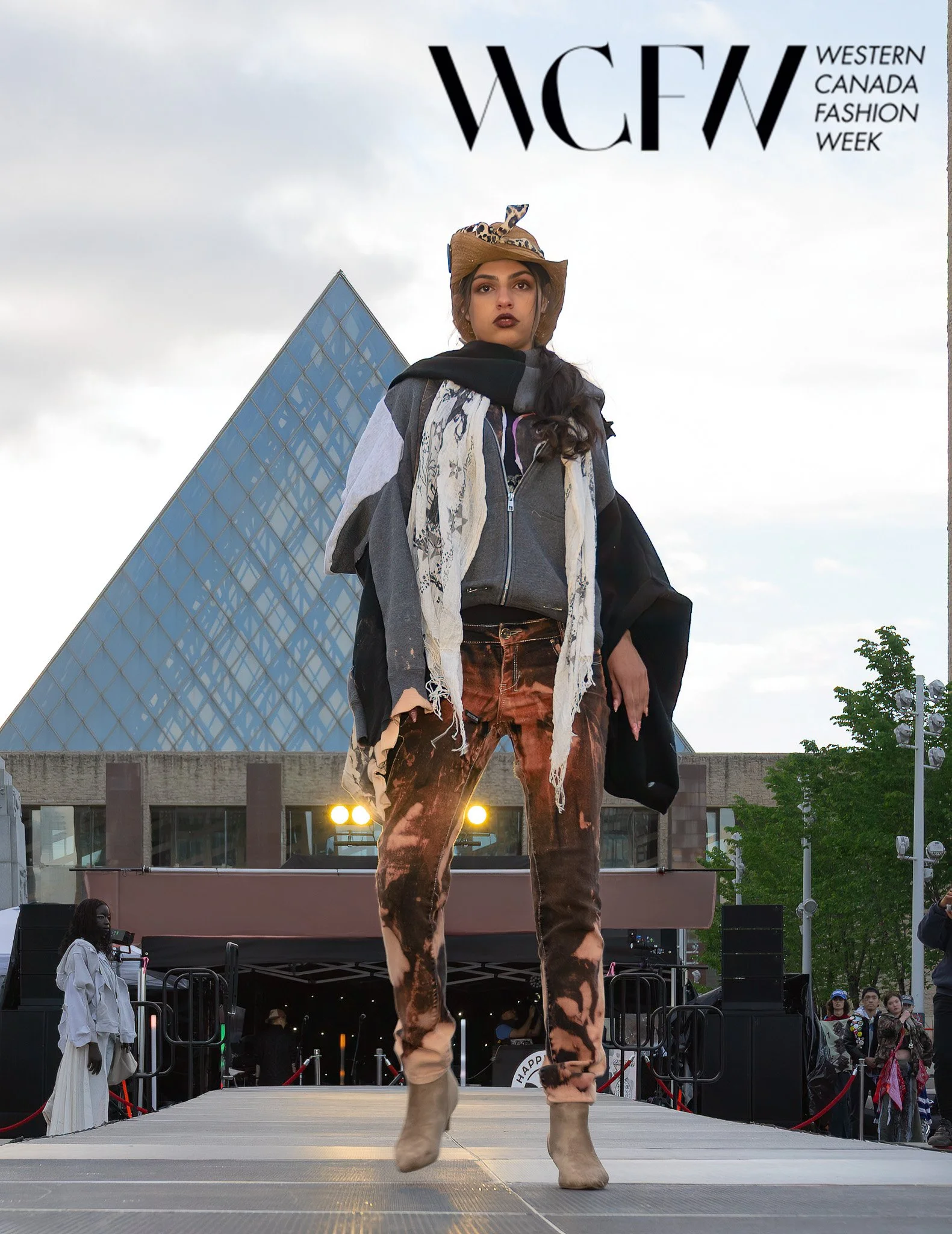 A woman walking on an outdoor stage during a fashion event, wearing a cowboy hat, gray jacket, and tie-dye pants, with a modern building and audience in the background.