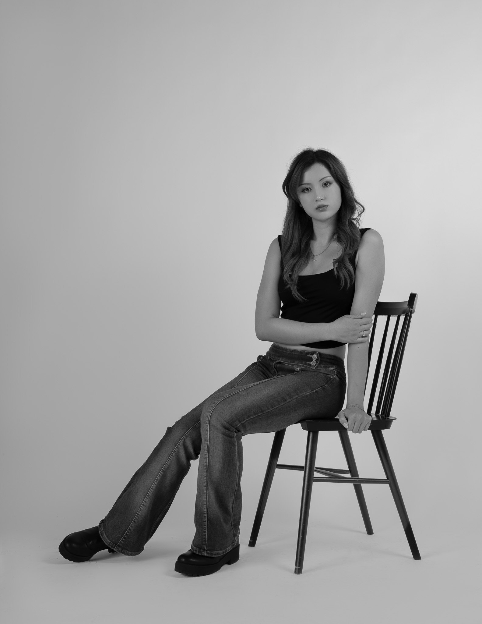 Black and white photo of a young woman sitting on a wooden chair against a plain background, wearing a black tank top and jeans with a confident expression.