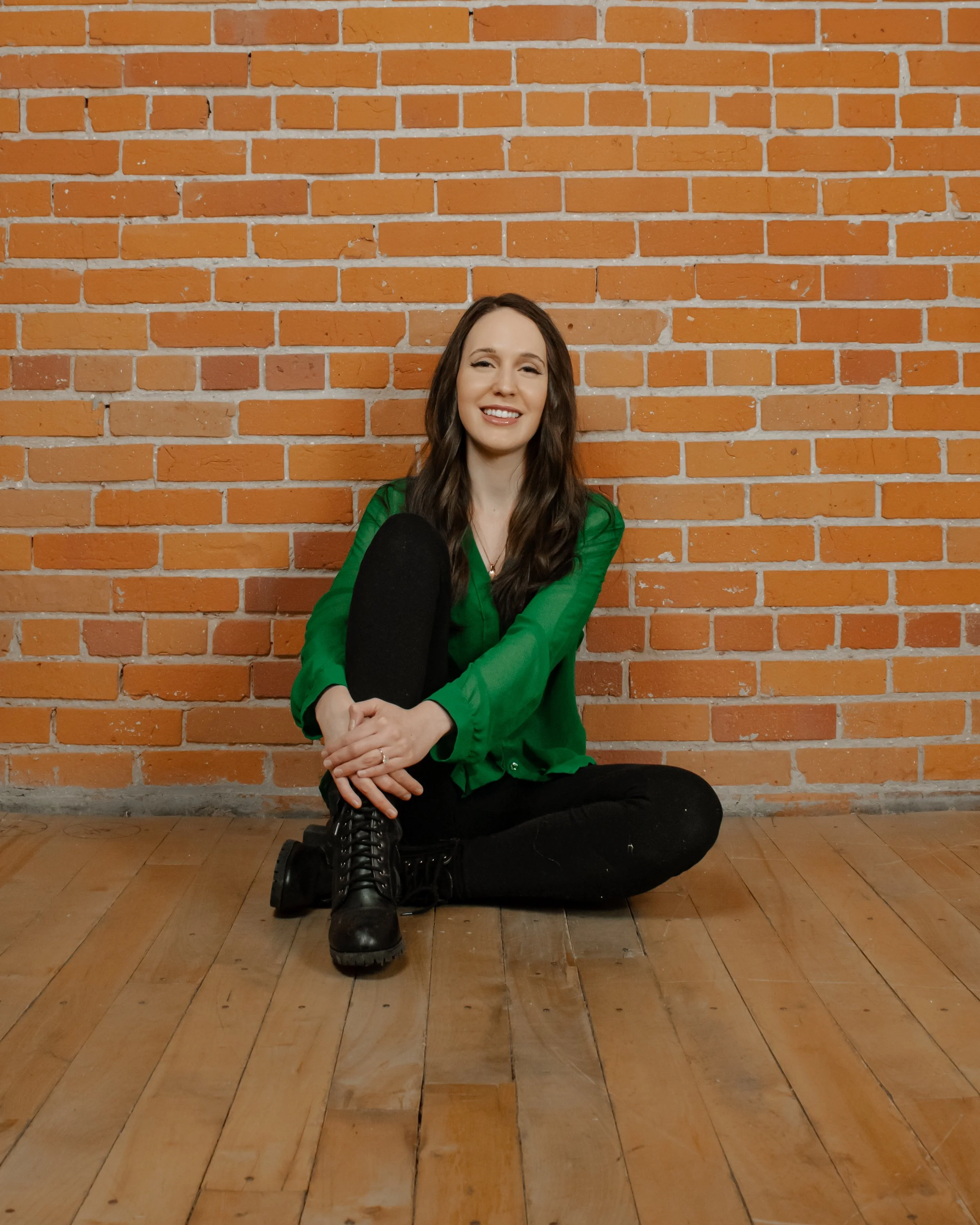 A woman with long dark hair sitting on a wooden floor in front of an orange brick wall, wearing a green blouse, black pants, and black boots, smiling and looking at the camera.