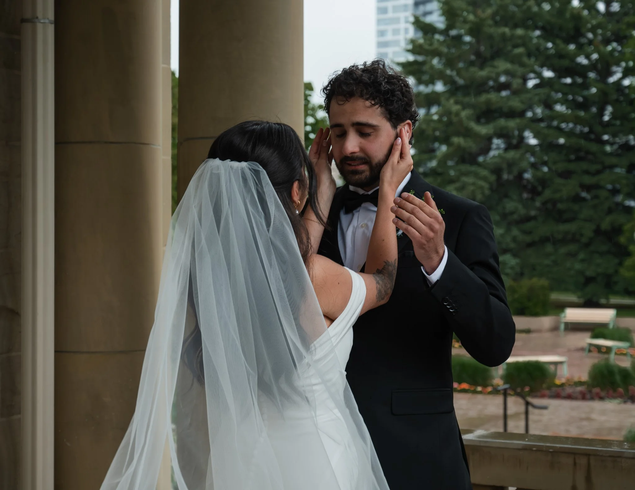 A bride and groom sharing a moment outdoors, with the bride gently touching the groom's face, both with closed eyes, near large stone columns and trees.
