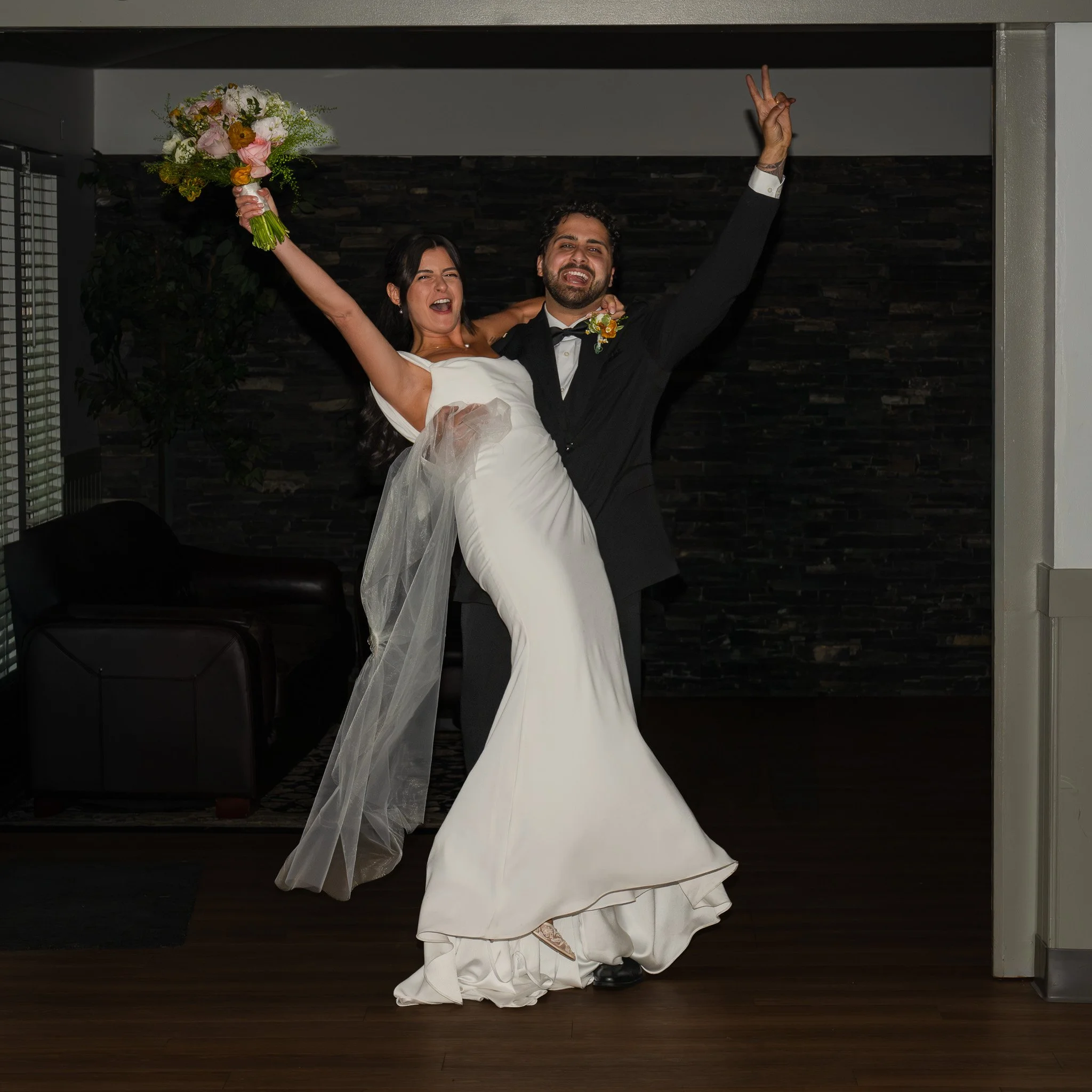 A bride holding a bouquet of flowers and a groom lift her in a celebratory pose. Both are smiling and dressed in wedding attire, with the bride in a white wedding dress and the groom in a black tuxedo.