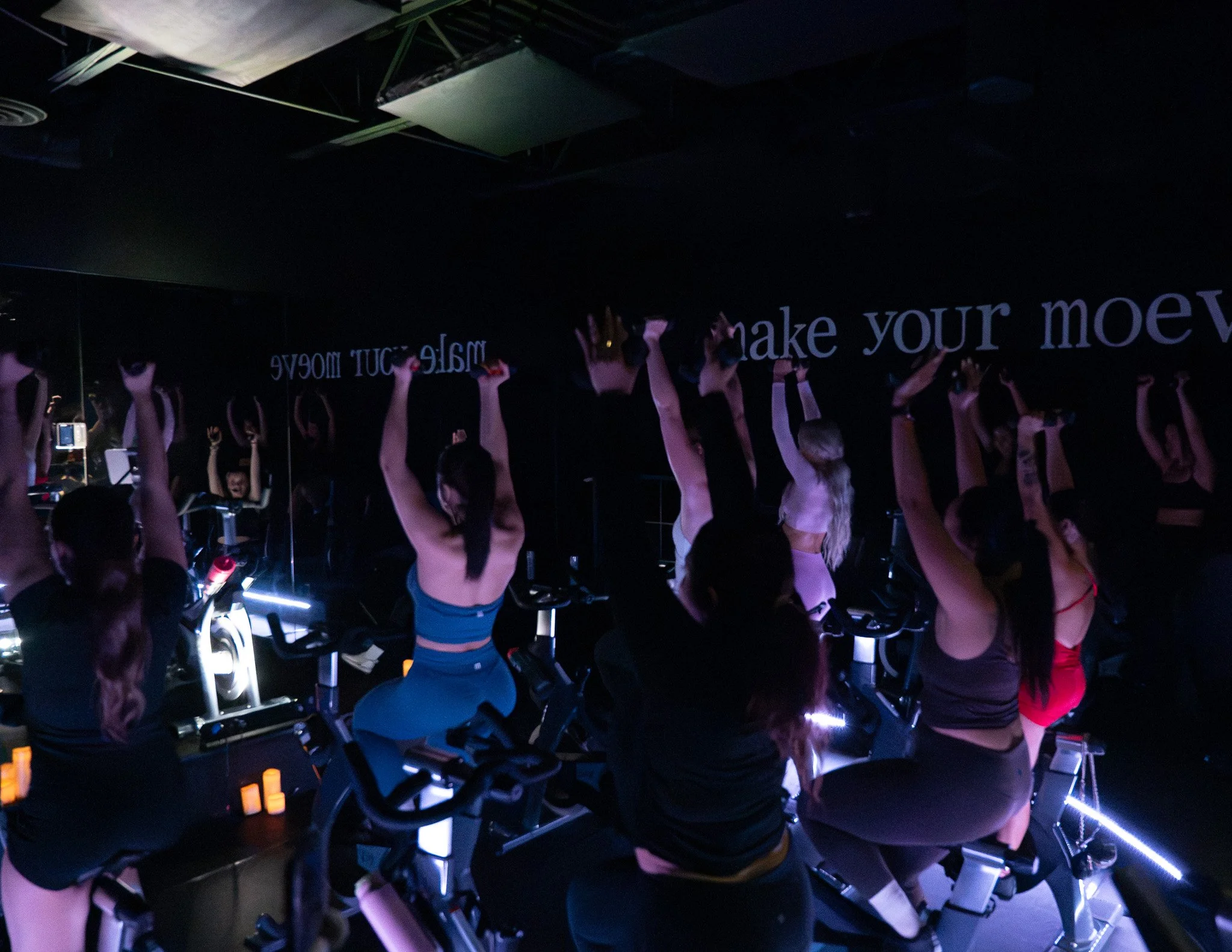 People participating in a fitness or yoga class in a dark room with neon lighting, some on stationary bicycles and others standing with their arms raised.