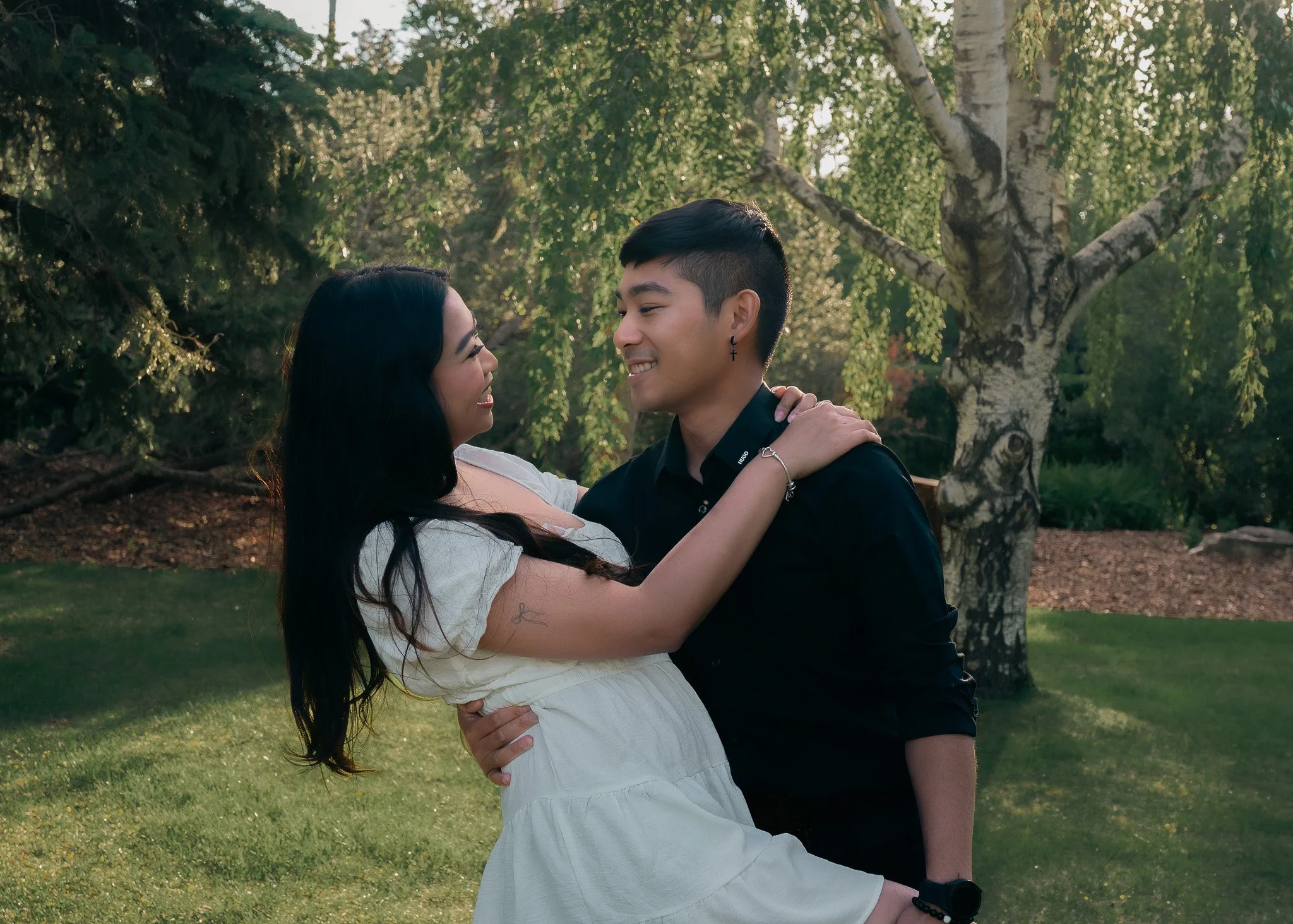 A young couple dancing outdoors in a park with green trees in the background. The woman has long dark hair, and the man has short dark hair and earrings. They are smiling and looking at each other affectionately.