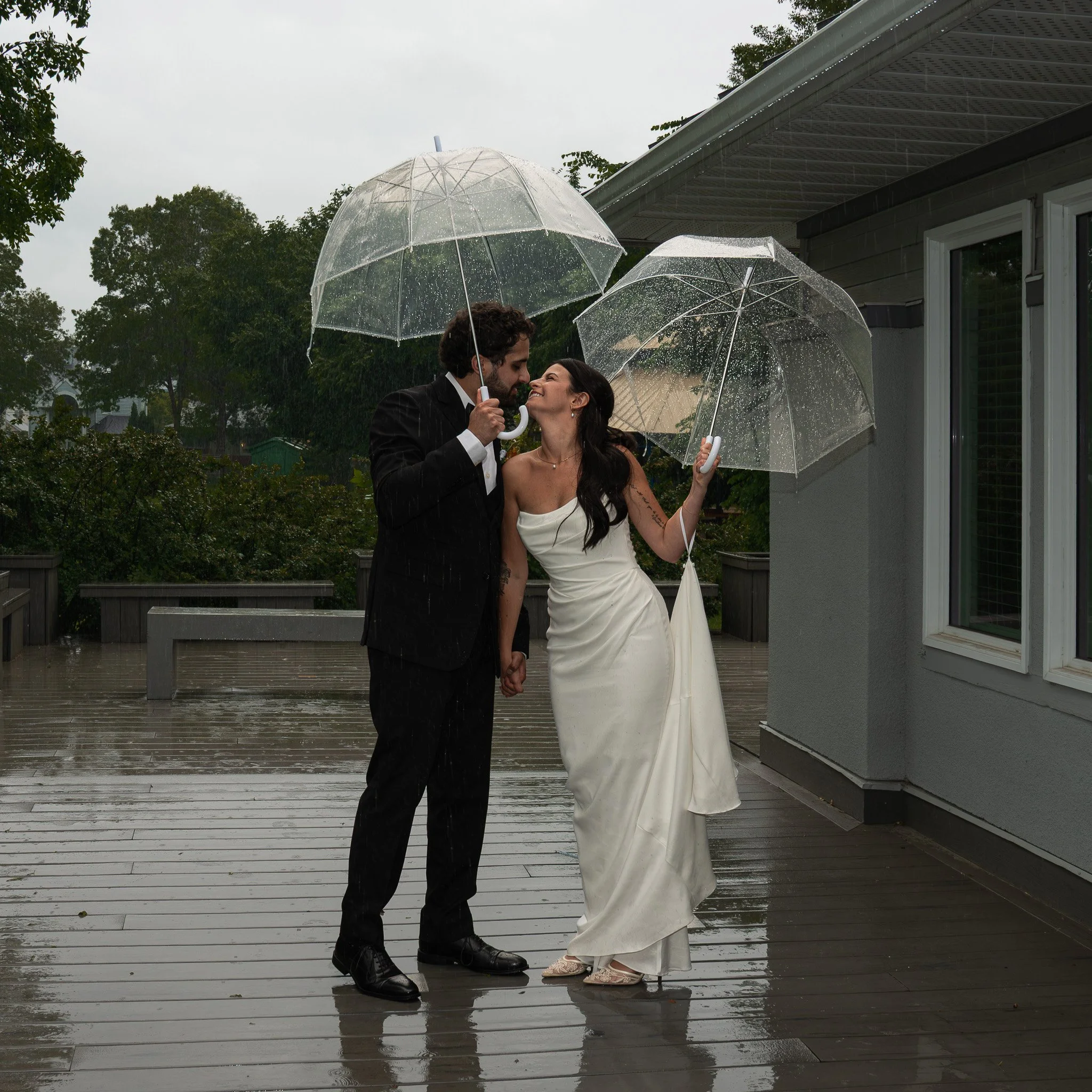 A bride and groom holding clear umbrellas in the rain, standing on a wooden deck outside a modern house with a gray siding. The groom wears a black suit and tie, and the bride wears a strapless white wedding dress. They are smiling and facing each ot