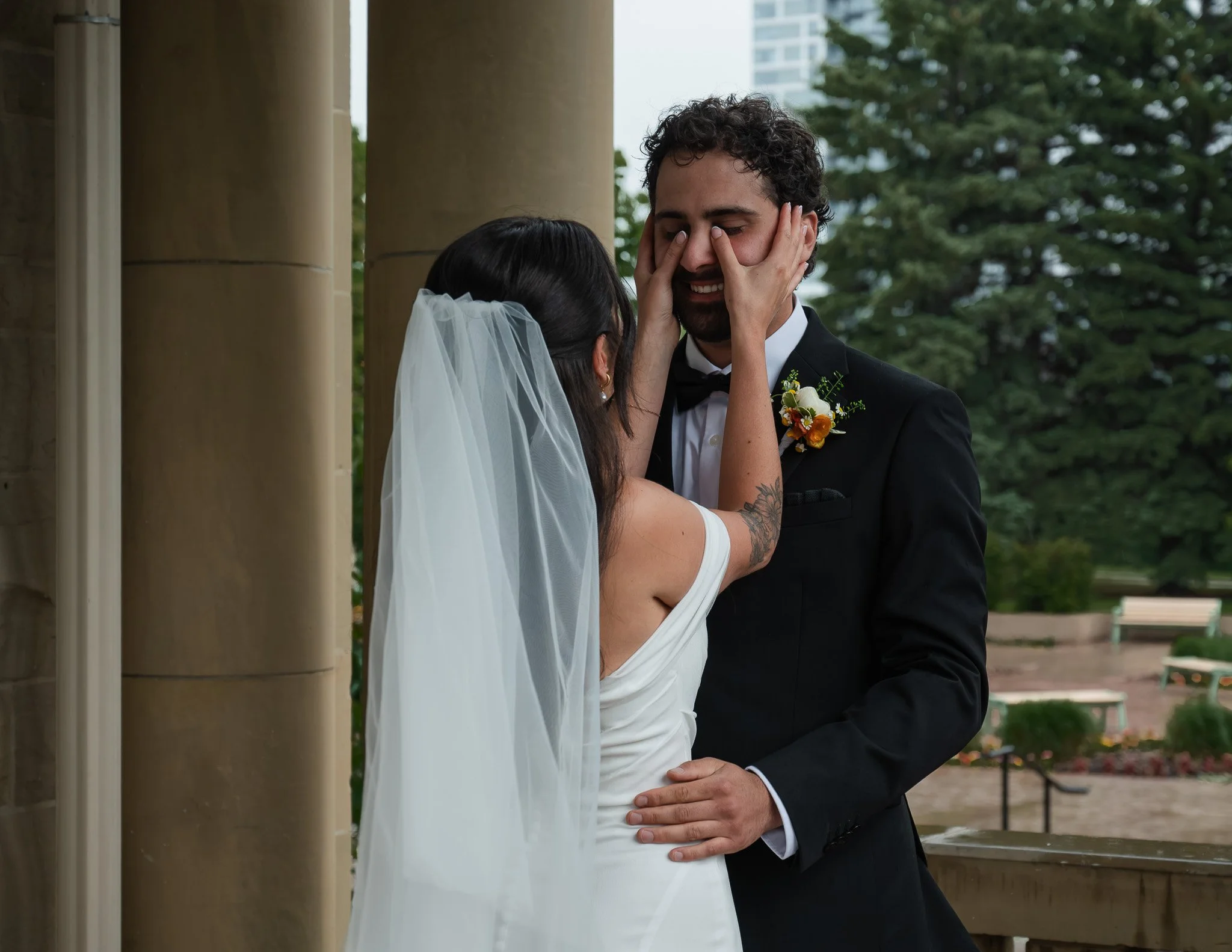 A bride and groom share an intimate moment outdoors during their wedding, with the bride gently holding the groom's face and the groom smiling while closing his eyes.