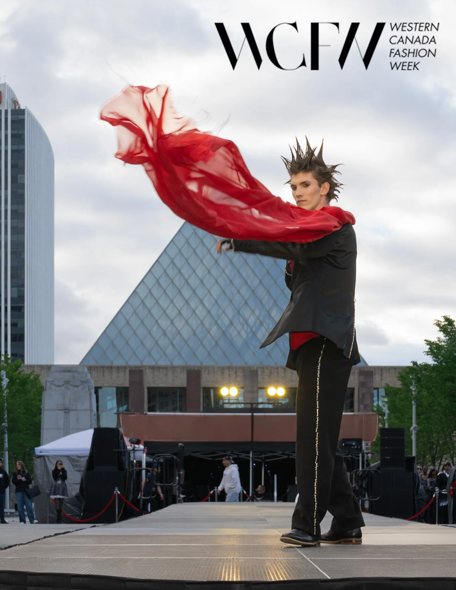 A person with styled hair, dressed in black and red, holding a flowing red fabric in an urban outdoor setting during cloudy weather, with a stage and people in the background at Western Canada Fashion Week.