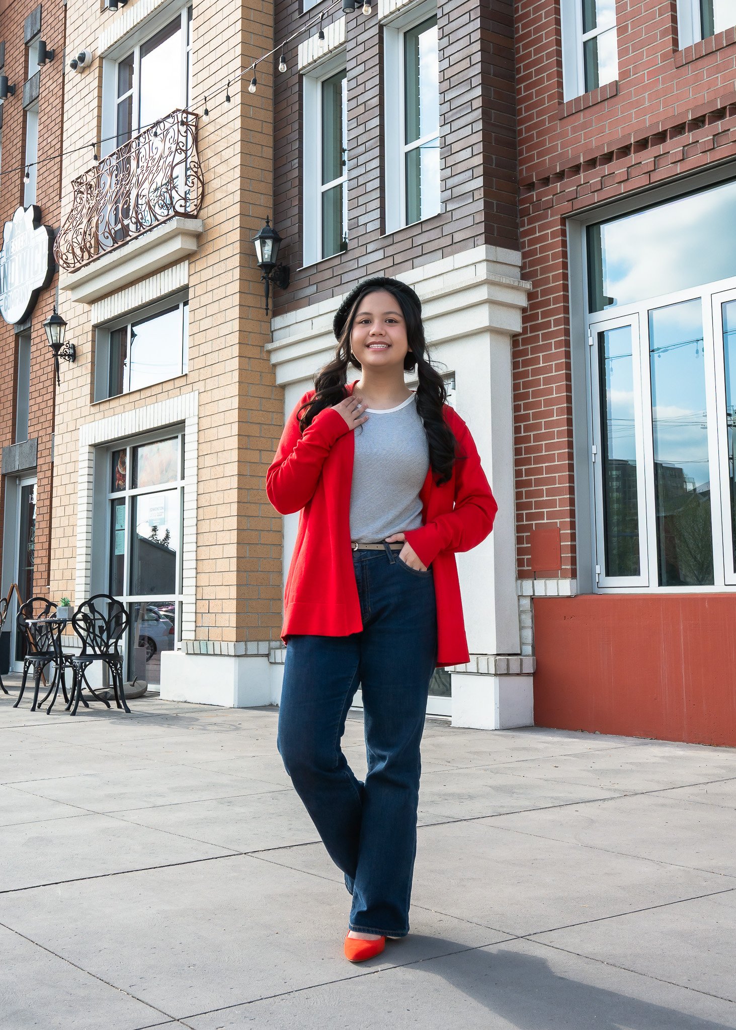 A young woman with long dark hair, smiling, standing on a sidewalk in front of a brick building with large windows. She is wearing a gray top, red coat, blue jeans, and orange shoes.