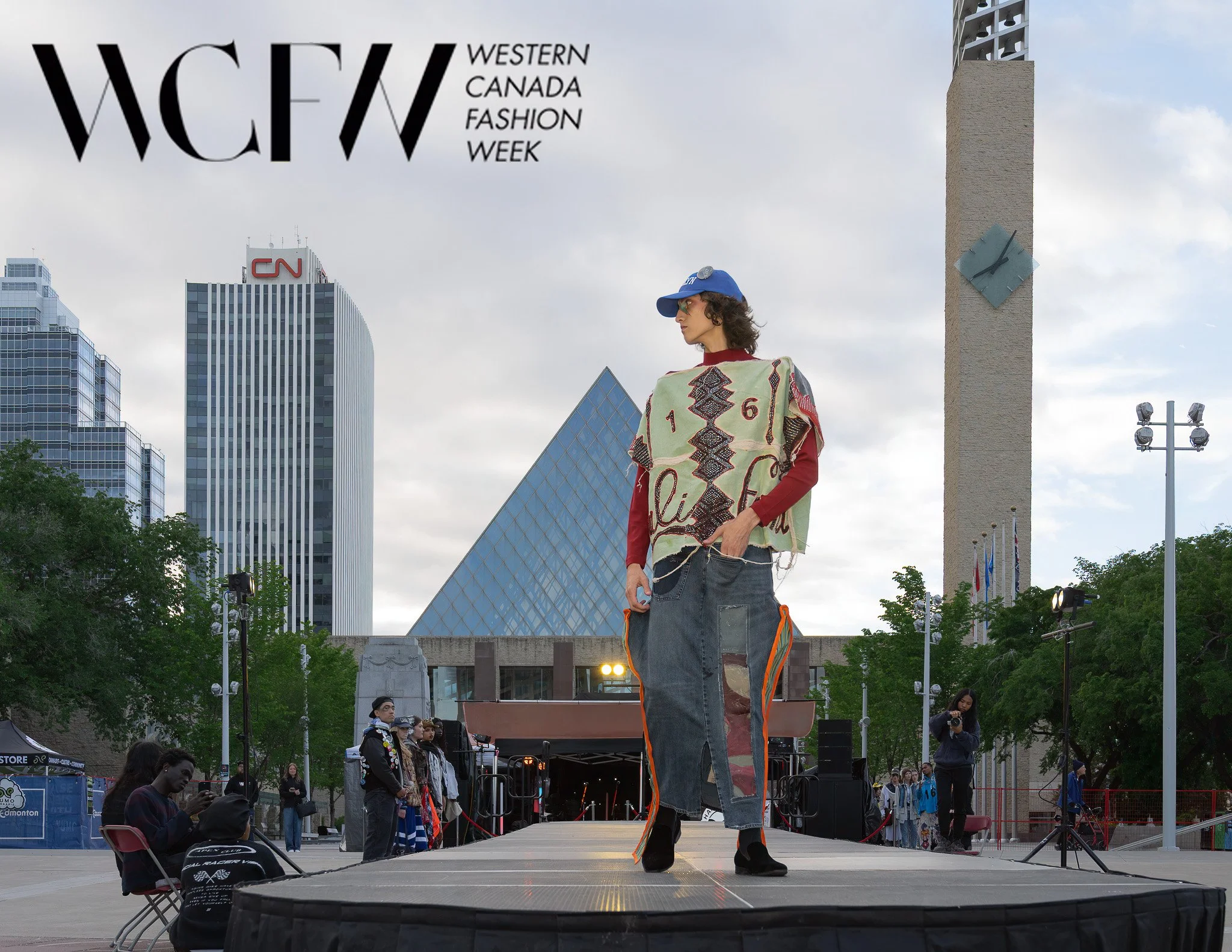 A model walking on stage during Western Canada Fashion Week in an urban outdoor setting, with tall modern buildings and a clock tower in the background, and audience members sitting on chairs in the foreground.