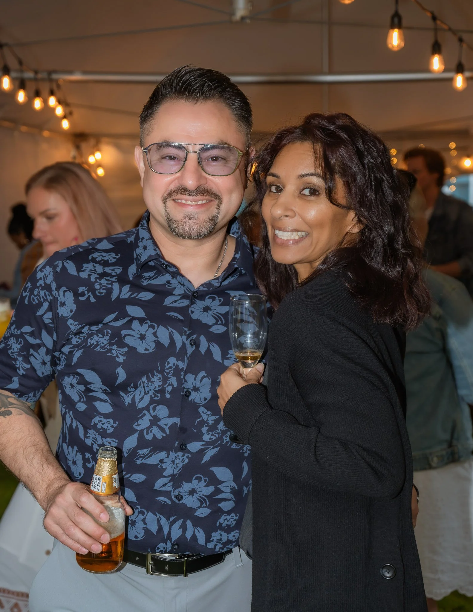 A couple smiling at an event, the man is holding a bottle of beer and the woman is holding a champagne flute. The background features warm string lights and other people.