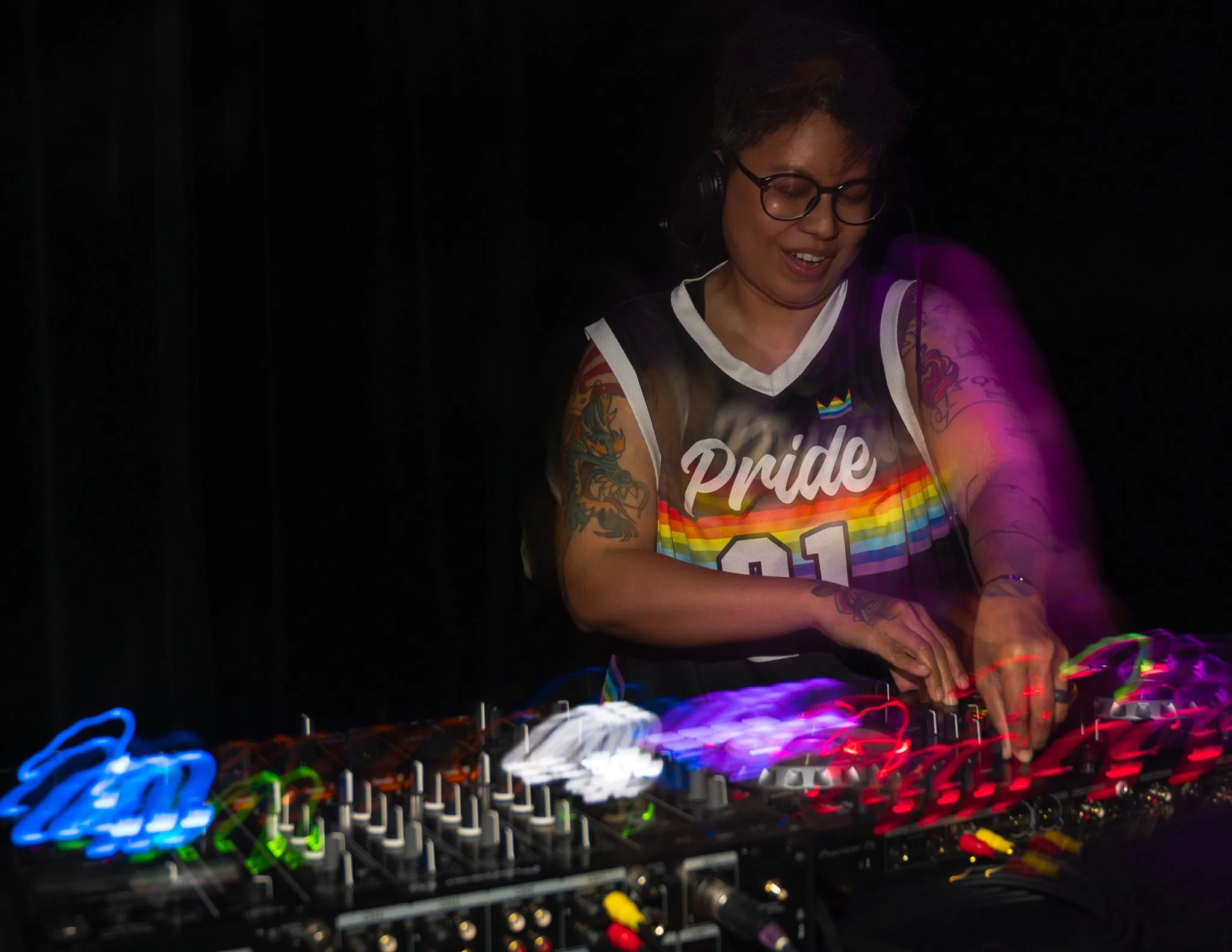 A person with short hair, glasses, and tattoos is DJing with turntables in a dark setting with colorful lights.