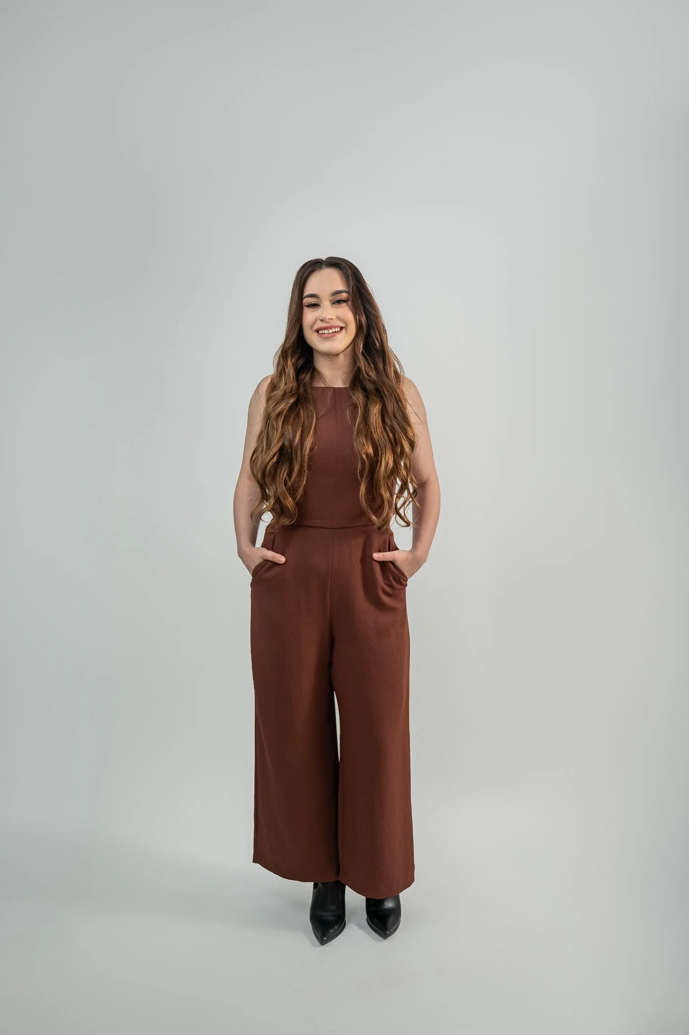 A young woman with long wavy brown hair, wearing a sleeveless brown jumpsuit, smiling with hands in pockets, standing against a plain light gray background.