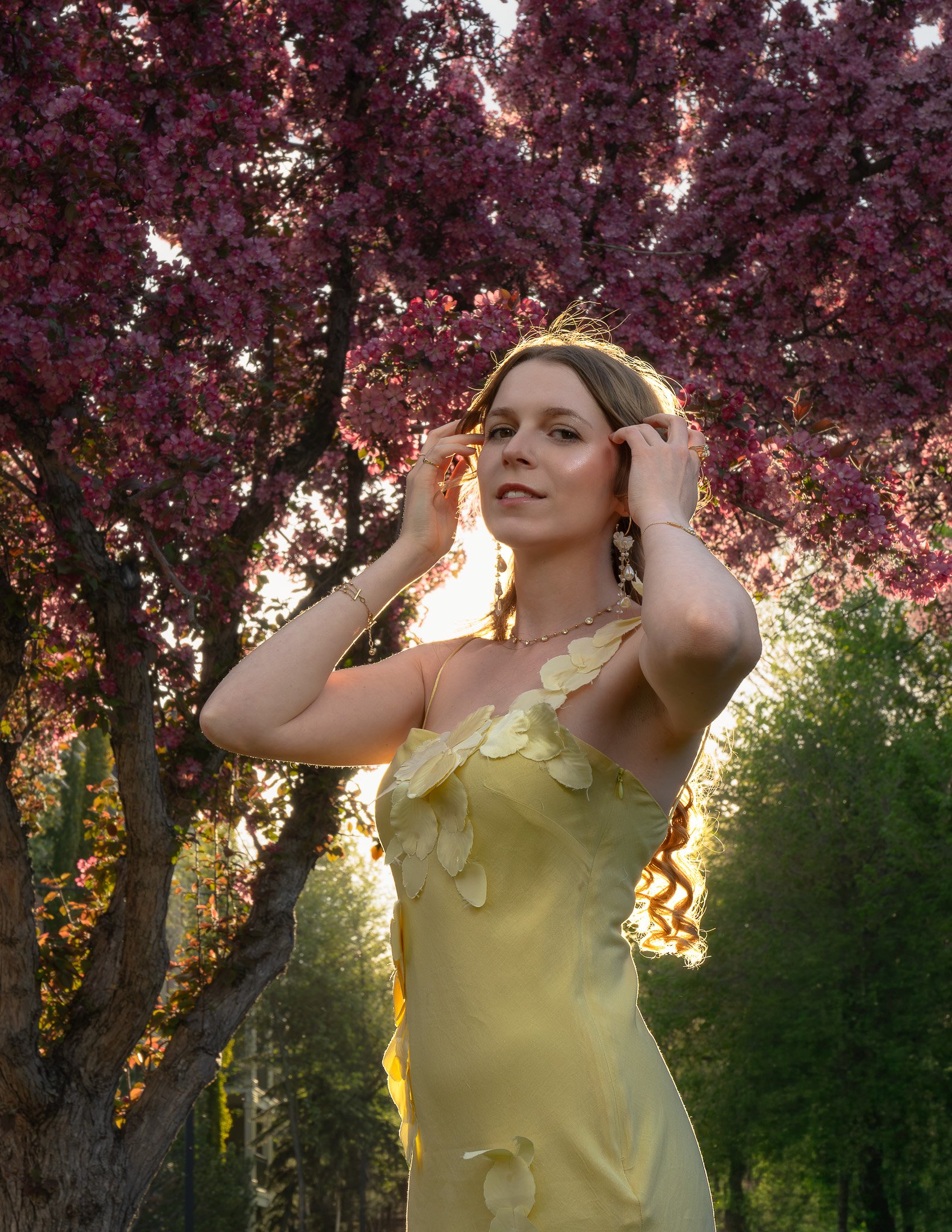 A woman in a yellow dress with flower details standing outside during sunset, with pink blossoms on a tree behind her and sunlight shining around her head.