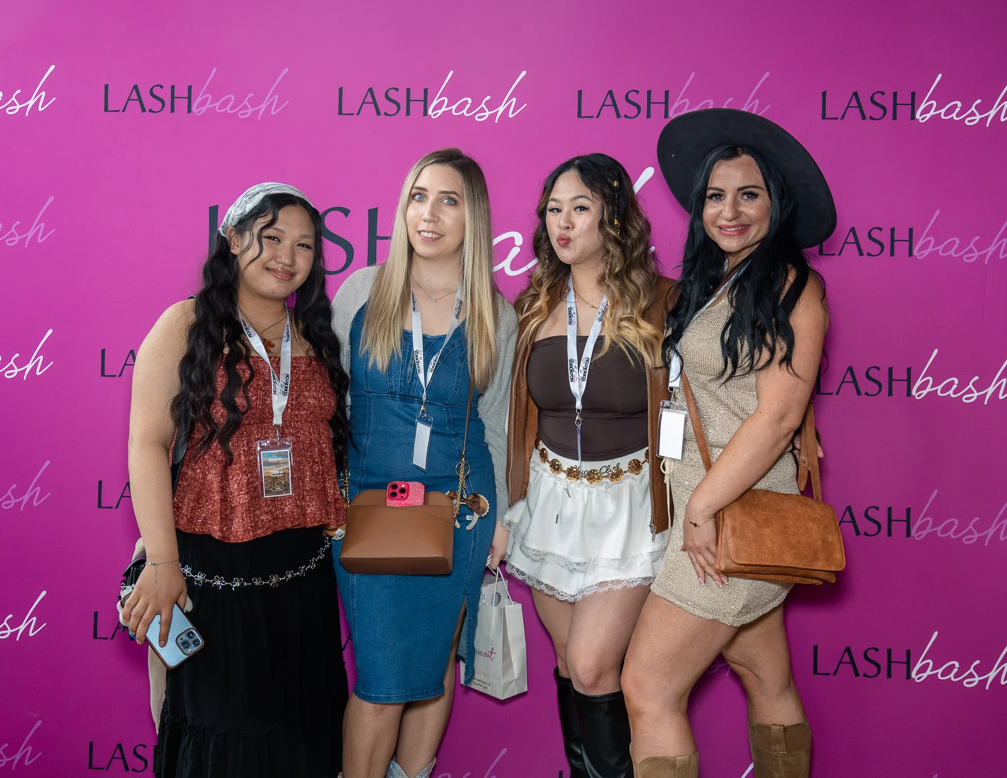 Four women standing together in front of a pink background with 'LASHbash' written repeatedly. They are smiling and dressed in fashionable clothing, some holding bags and a phone.