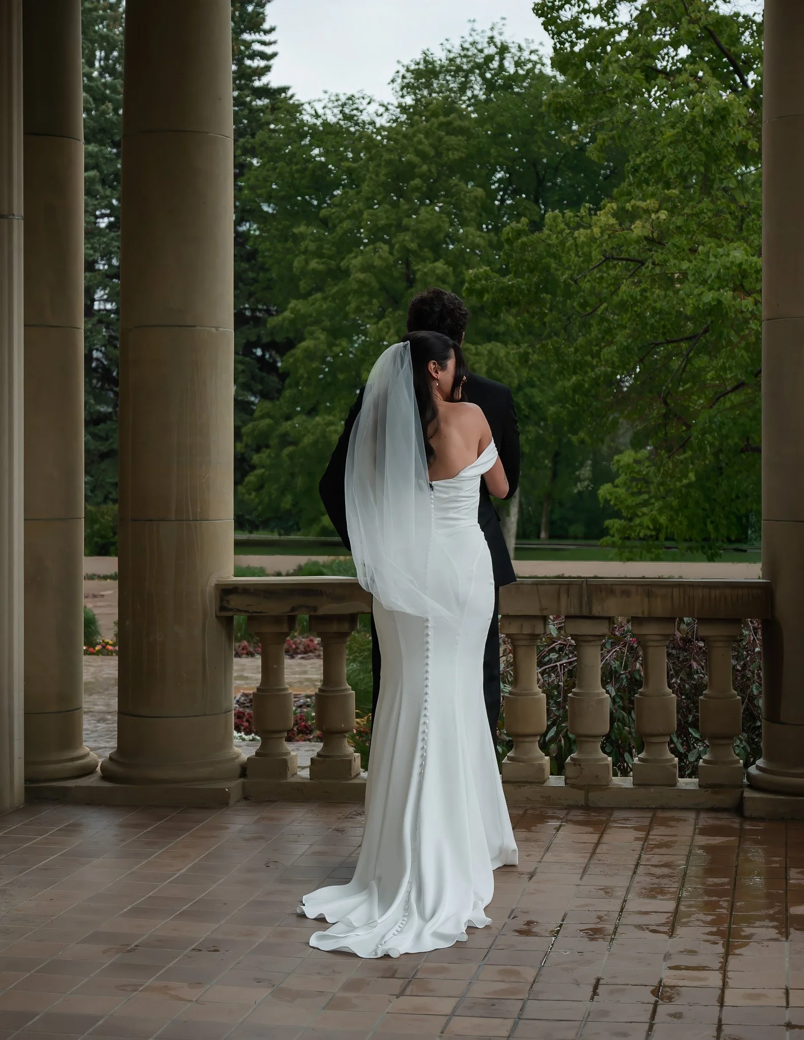 A bride in a white wedding dress and veil standing next to a groom in a black suit on a porch with tall columns, overlooking a garden with green trees, during rainy weather.