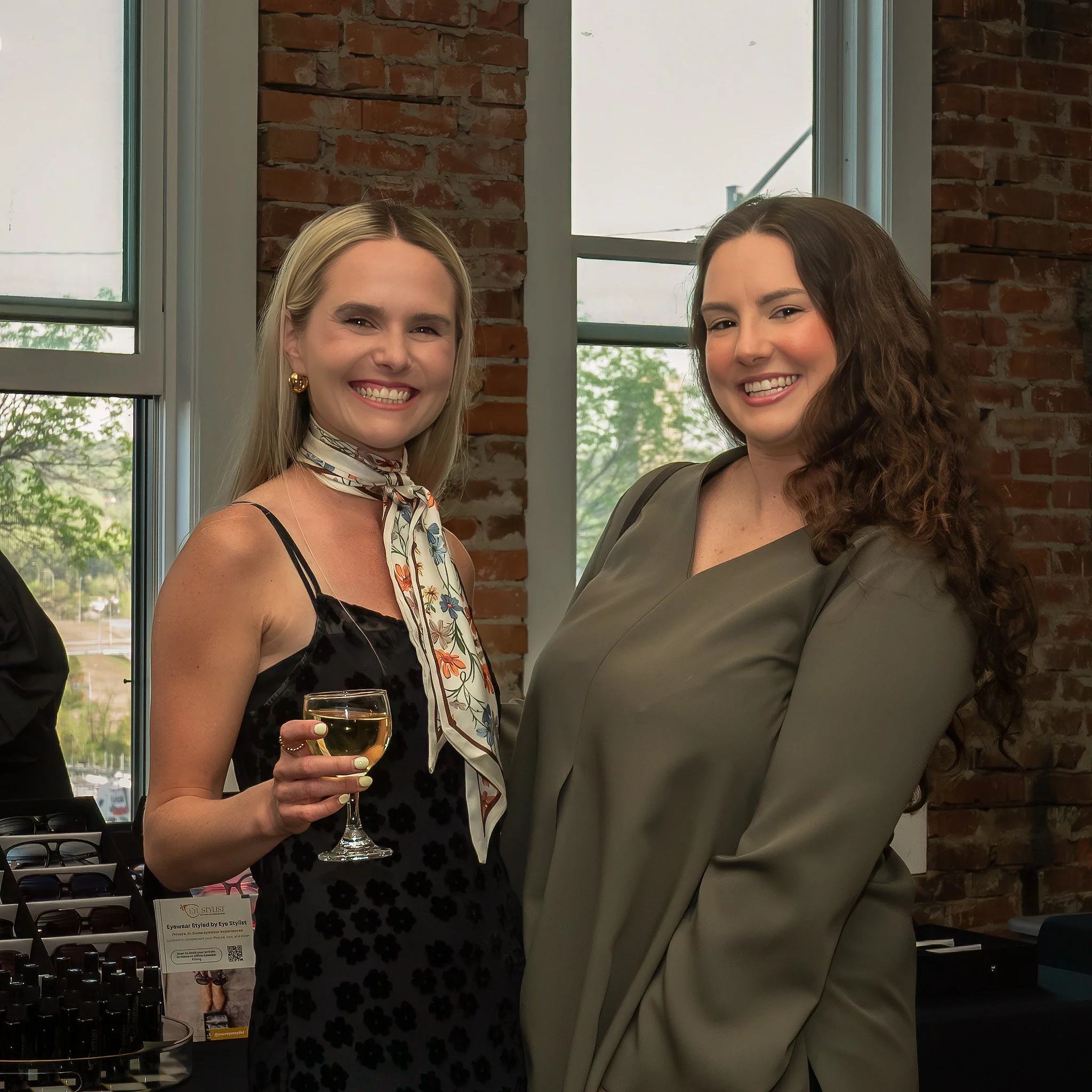 Two women smiling and posing for a photo indoors near large windows. One woman is holding a glass of white wine, and a display of eyeglasses is visible in the foreground.