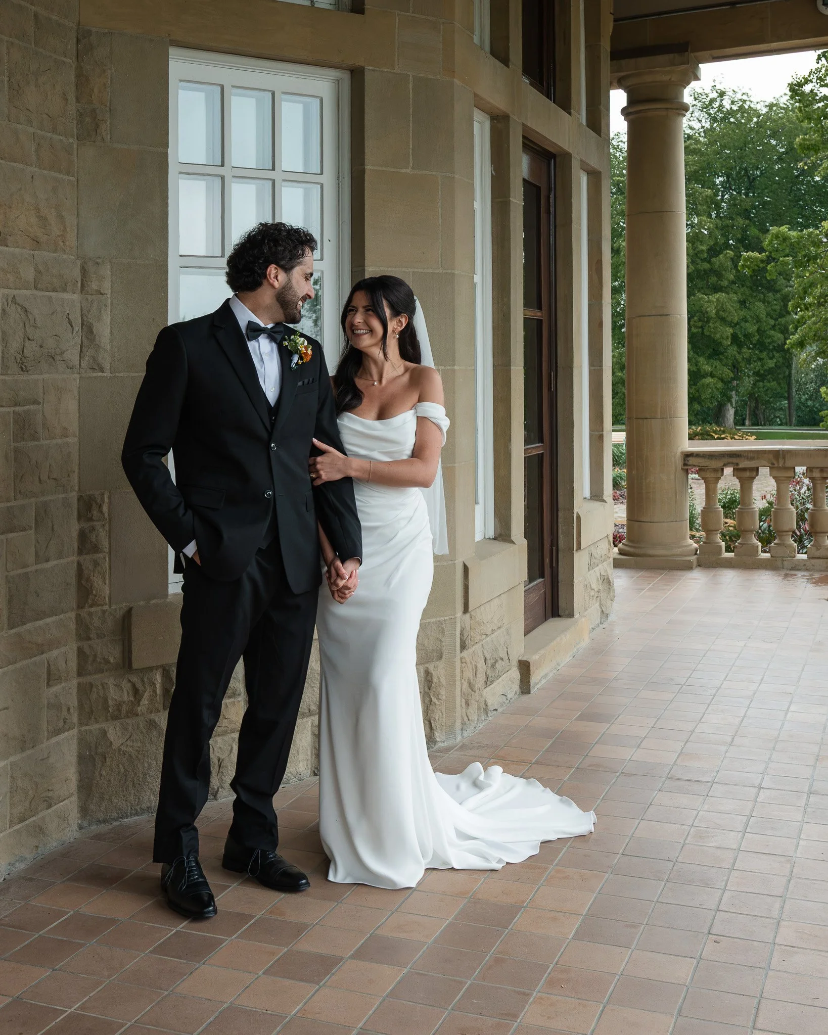 A bride and groom standing on a brick patio outside a stone building, smiling and looking at each other.