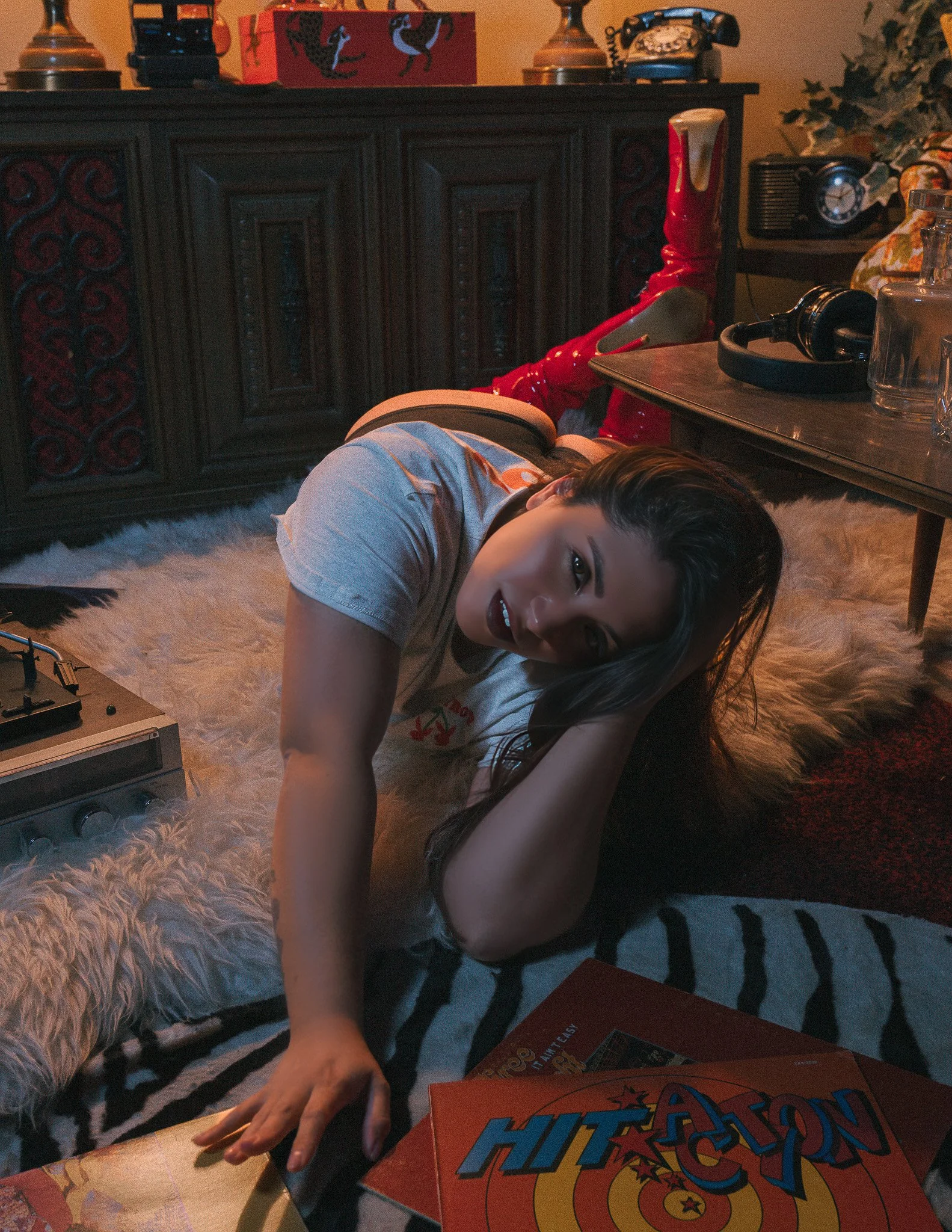A young woman lying on the floor next to a table, smiling at the camera. Behind her is a dark wooden cabinet, and on the table are various items including a board game titled 'Hit Action,' a pair of headphones, a clear bottle, and other objects. The 