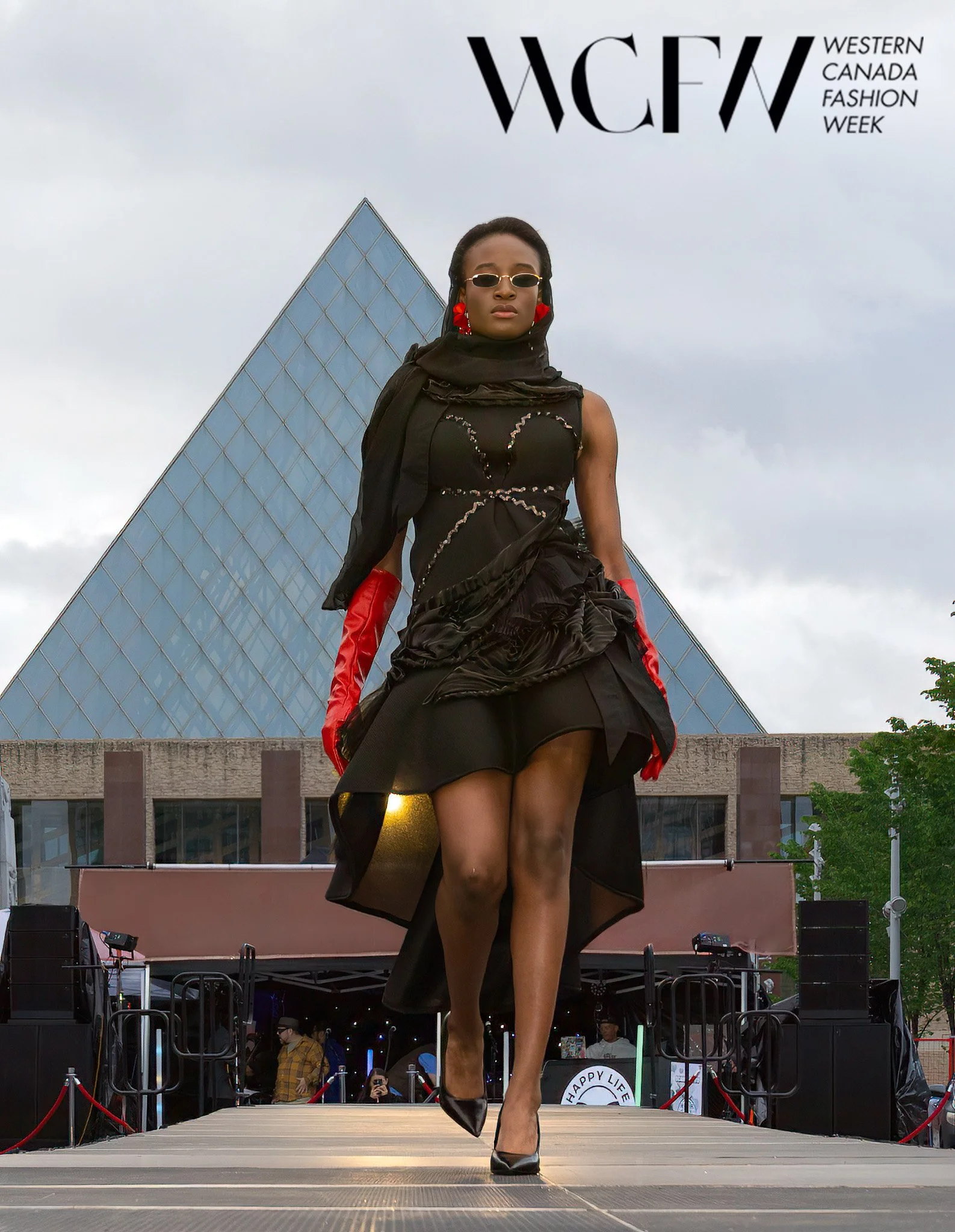 A fashion model walking down a runway at Western Canada Fashion Week, wearing black and red clothing, sunglasses, and red gloves.