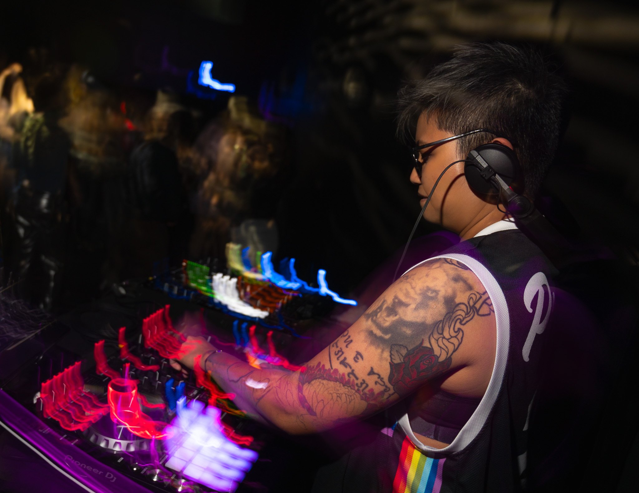 A DJ with short dark hair, glasses, and tattoos on their arms, wearing a black and white vest with a rainbow stripe on the shoulder, and headphones over their ears, performing at a dark party with colorful, blurred lights and DJ equipment.