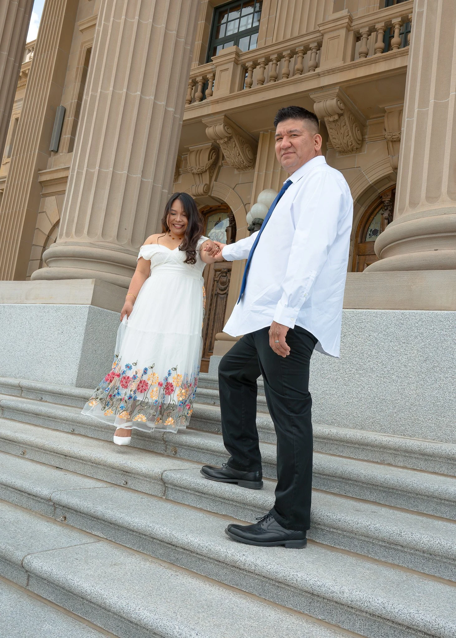 A man in a white shirt and black pants holding hands with a woman in a white dress with floral embroidery, standing on steps outside a grand building with large columns and ornate architectural details.