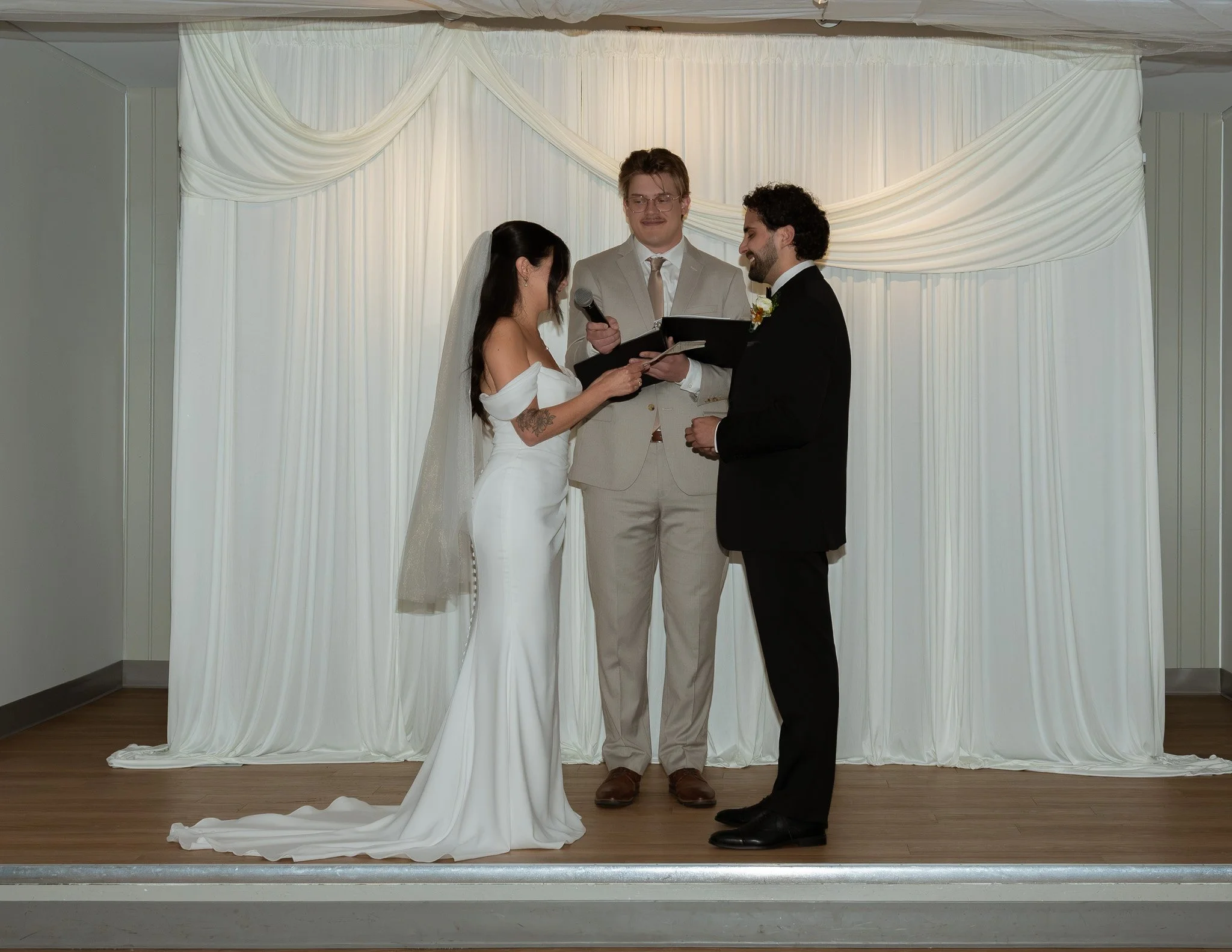A couple getting married during a wedding ceremony, with a man reading wedding vows from a book, and the bride holding his hand, under white draped fabric in the background.