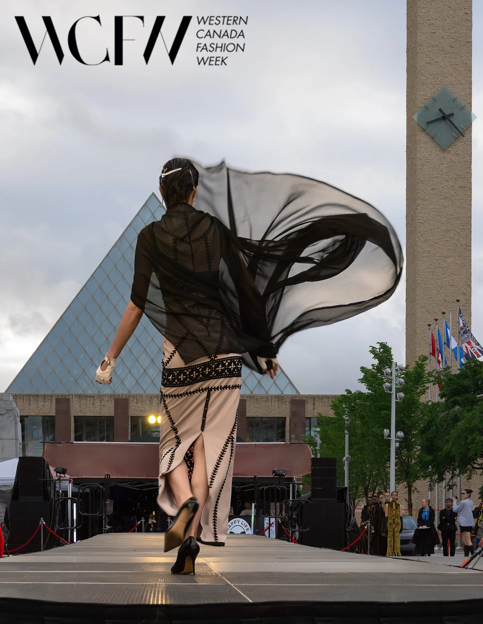 A model walking on an outdoor runway at Western Canada Fashion Week, wearing a black sheer top and beige pants with black embroidery, with a large black flowing scarf, against a backdrop of buildings, a clock tower, and people watching.