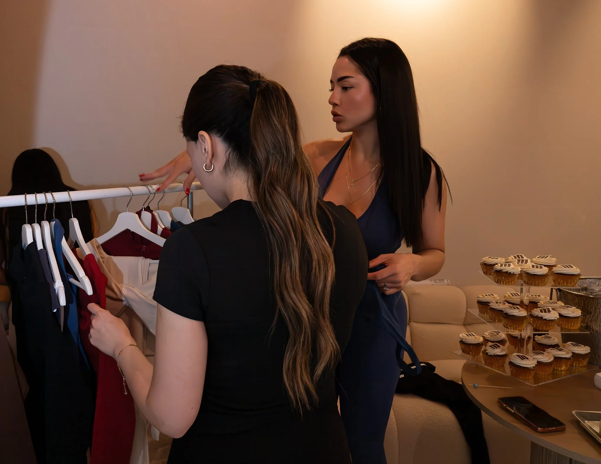 Two women looking at clothing on a rack in a boutique store, with desserts on a table in the background.