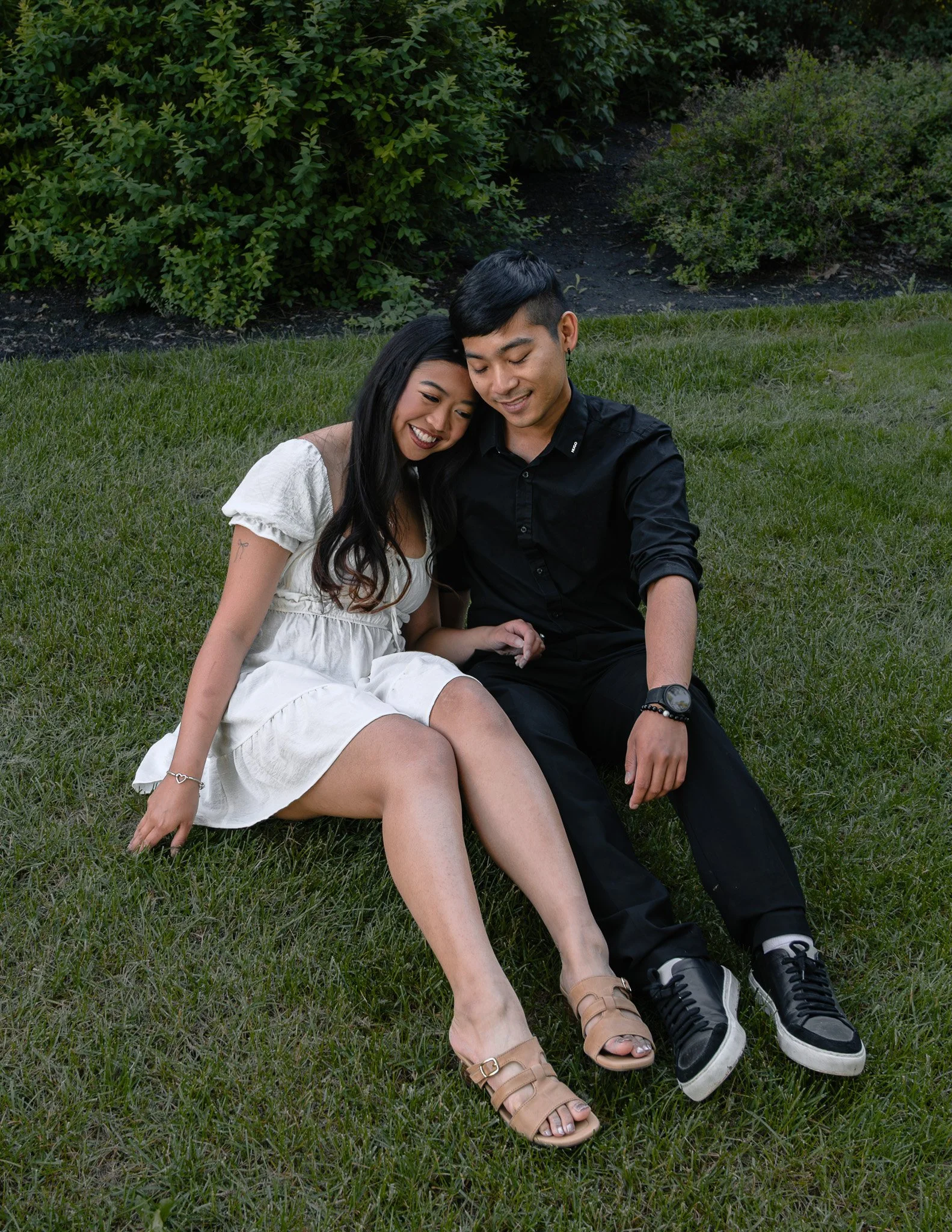 A young couple sitting closely together on the grass in a park, smiling and enjoying each other's company, with greenery and bushes in the background.