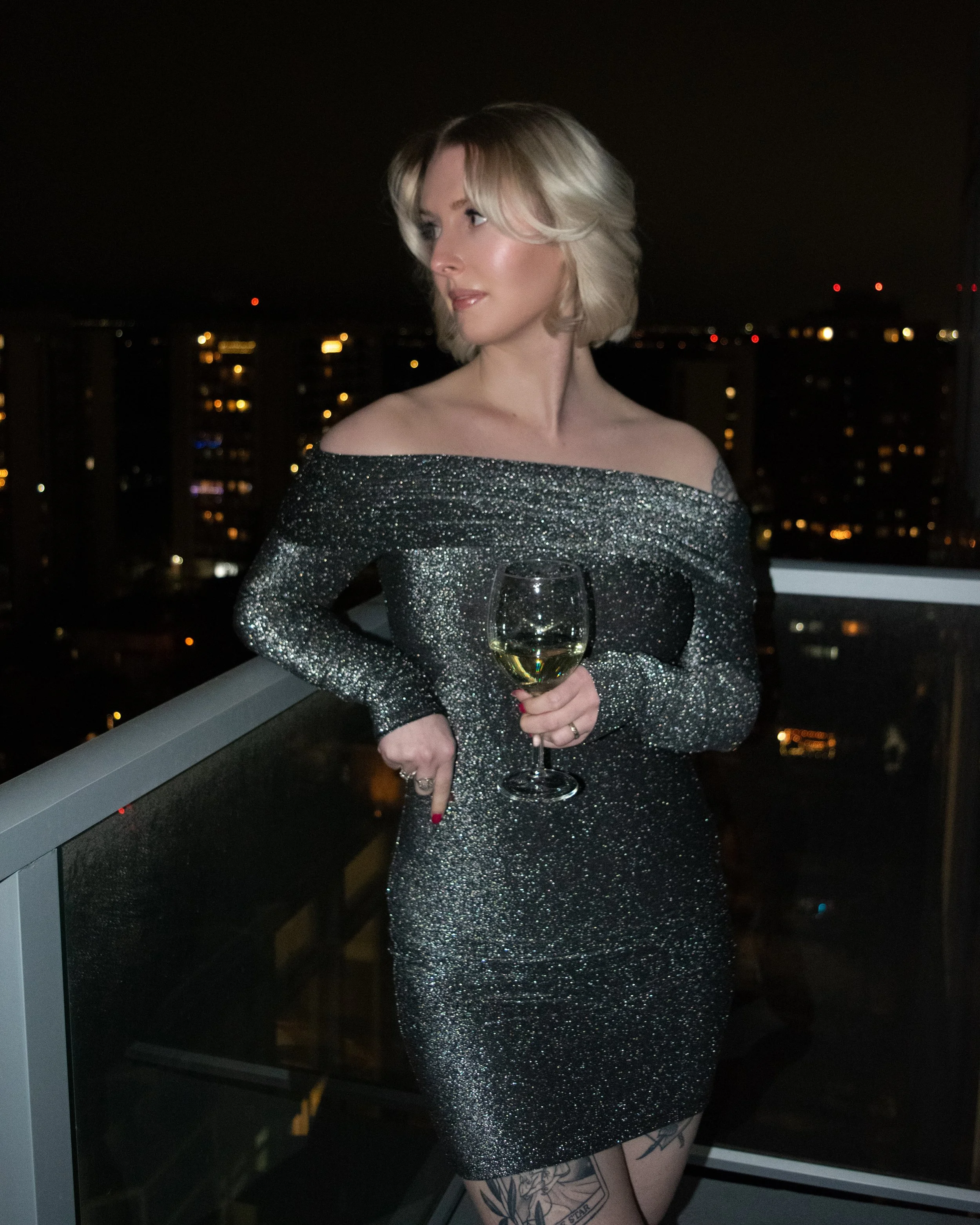 A woman with blonde, wavy hair in a sparkly off-the-shoulder dress holding a glass of white wine on a balcony at night with city lights in the background in Edmonton, Alberta.