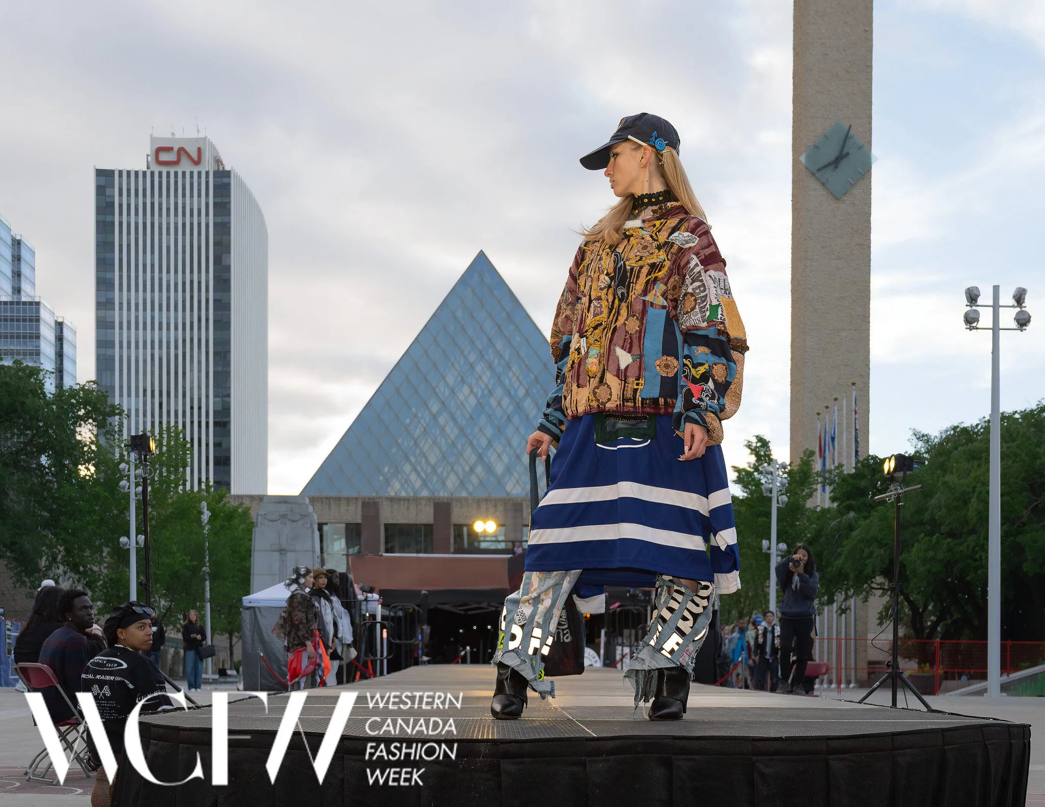 A woman models on a runway during Western Canada Fashion Week in an urban park setting with skyscrapers and cloudy sky in the background. She wears a colorful patchwork jacket, wide-leg pants with bold text, and black boots. Audience members and phot