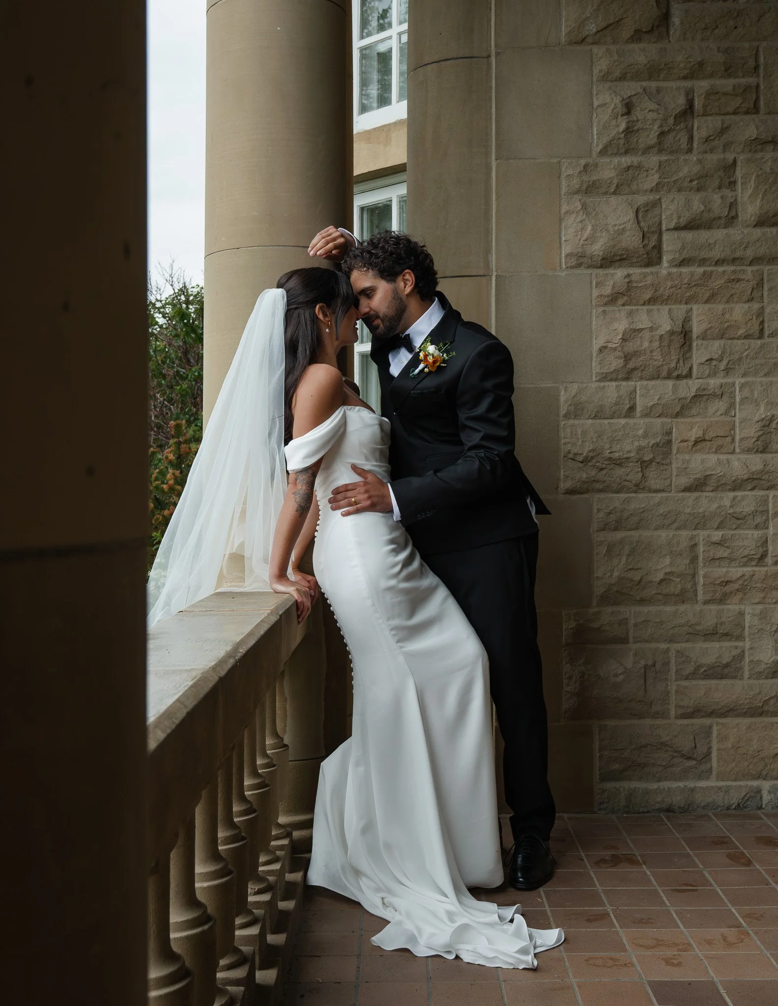 A bride and groom are standing close together, forehead to forehead, on a porch or balcony with stone columns. The bride is wearing a white wedding gown with a veil, and the groom is in a tuxedo with a boutonniere. They appear to be sharing an intima