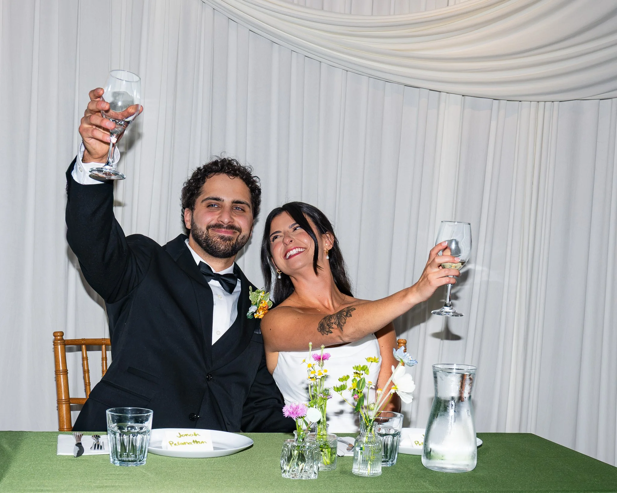 A man in a tuxedo and woman in a white dress celebrating at a wedding reception, holding up wine glasses and smiling. They sit at a table decorated with flowers, with a snowy white curtain backdrop.