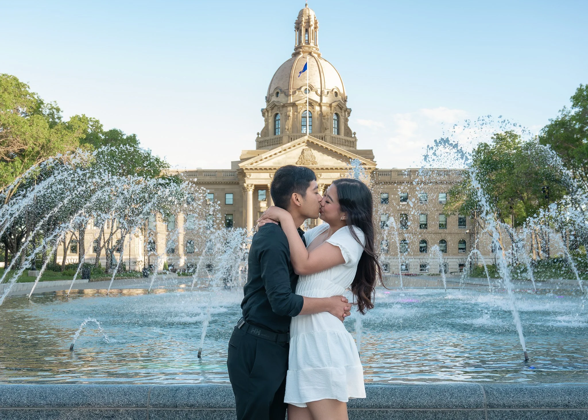 A couple kissing in front of a fountain with a large government building in the background during daytime.