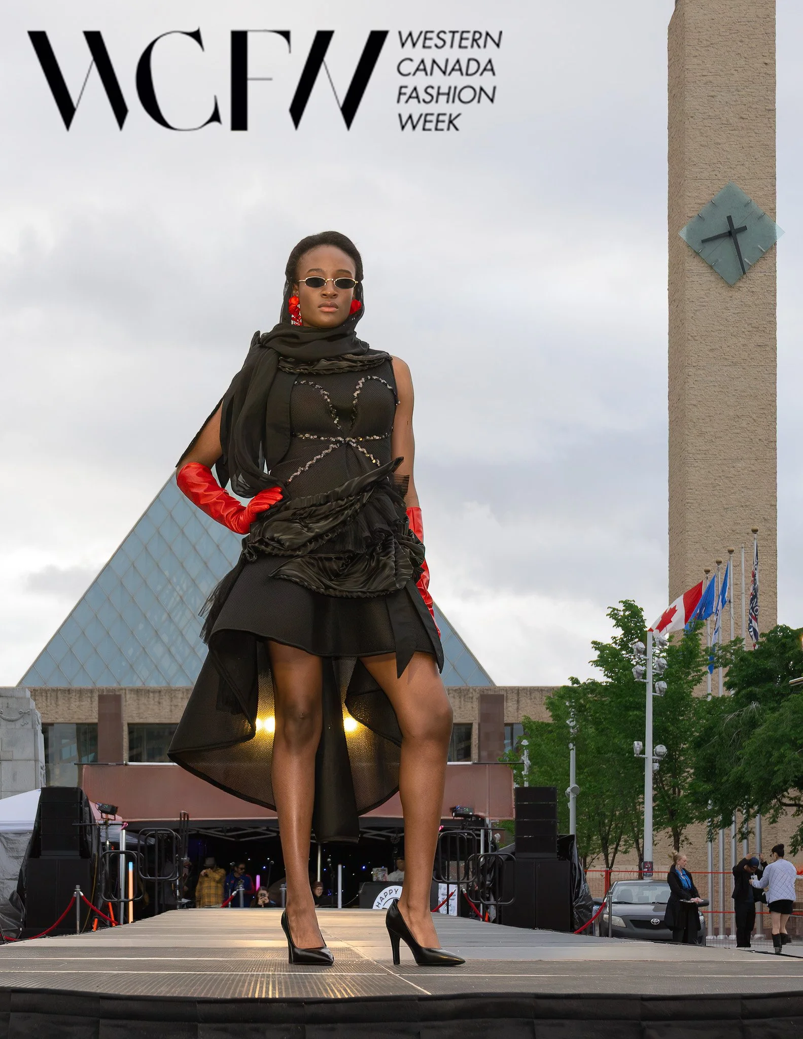 A woman walking on a stage at Western Canada Fashion Week, wearing black high heels, a black sheer skirt, a black top, red gloves, and sunglasses, with a building and flags in the background.