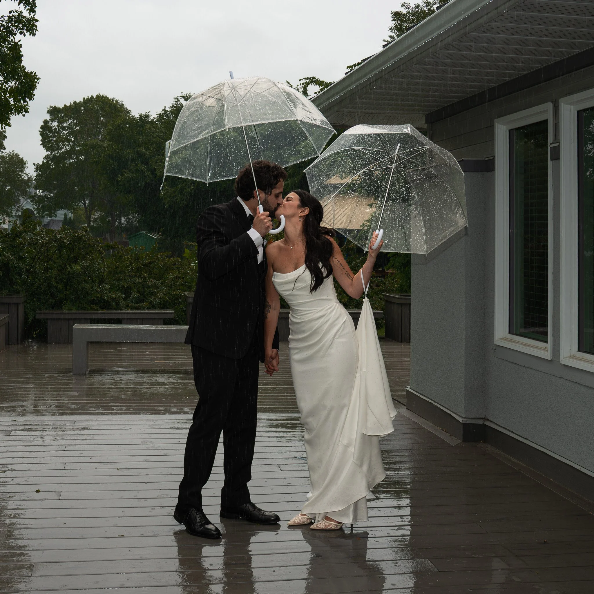 A couple sharing a kiss in the rain, holding clear umbrellas, standing on a wooden deck outside a house, with a woman in a white wedding dress and a man in a dark suit.