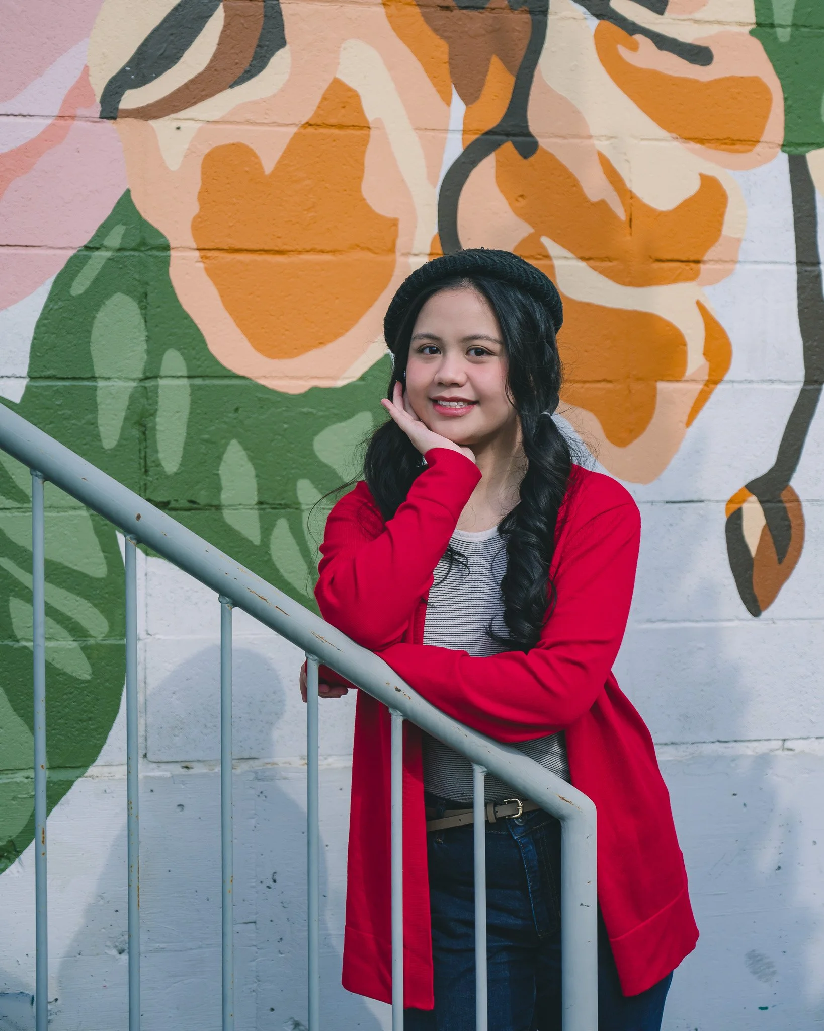 A young woman with long black hair, wearing a black beanie, a red coat, striped shirt, and jeans, standing outdoors in front of a colorful mural with large abstract orange, green, and beige shapes and smiley face outlines, smiling and posing with her