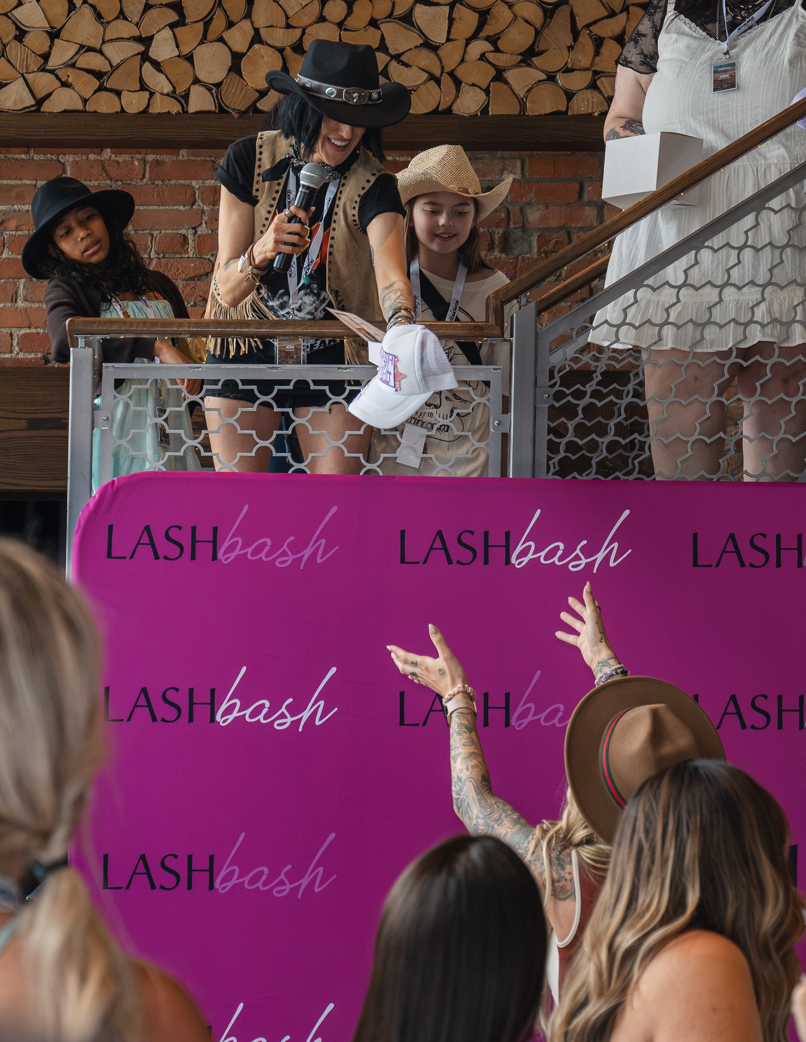 Woman in a cowboy hat and fringed vest signing a white cap for a fan at Lash Bash event, with several onlookers and a brick wall background.
