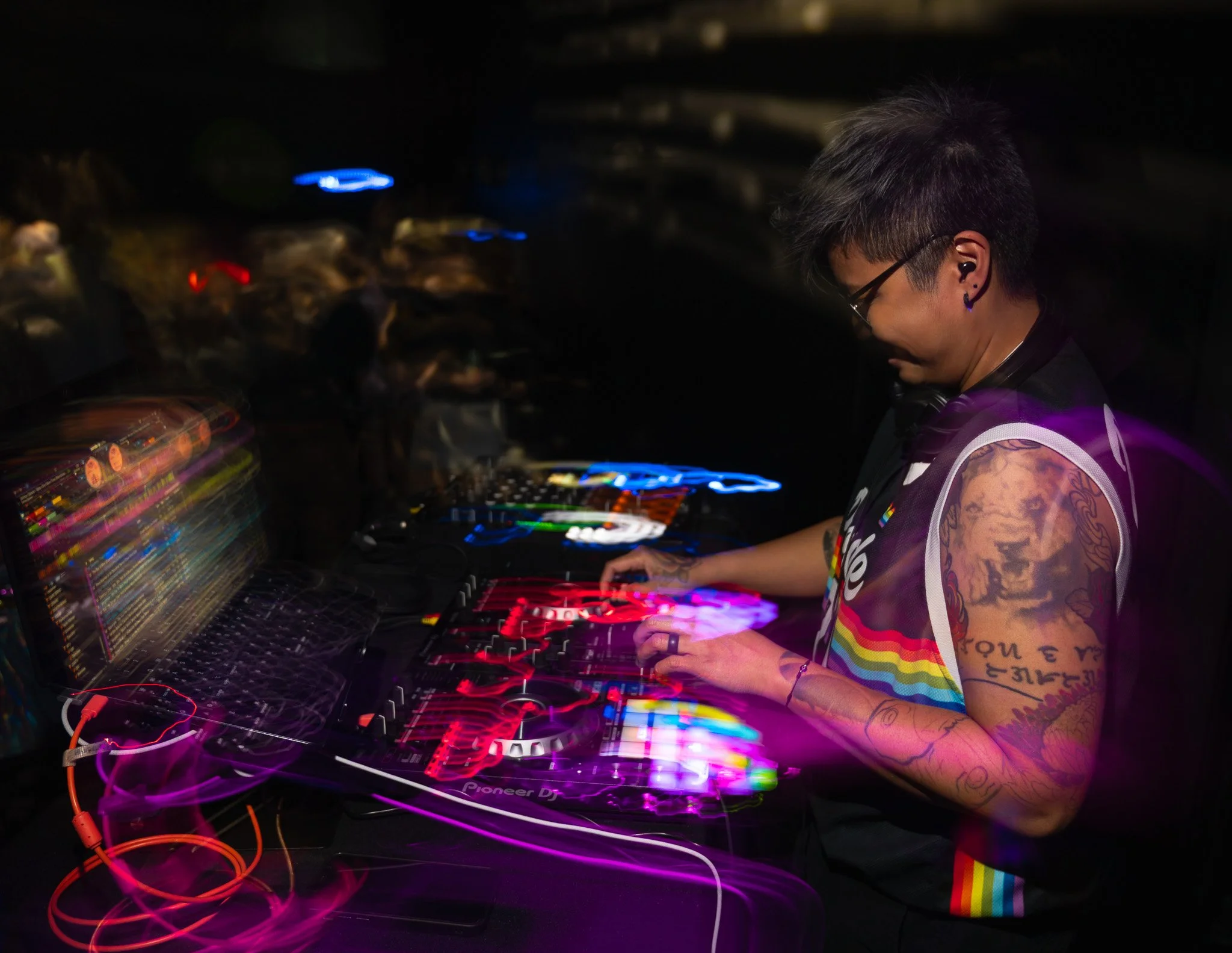 A person with tattoos and rainbow-colored attire DJing with colorful lights and equipment in a dark setting.