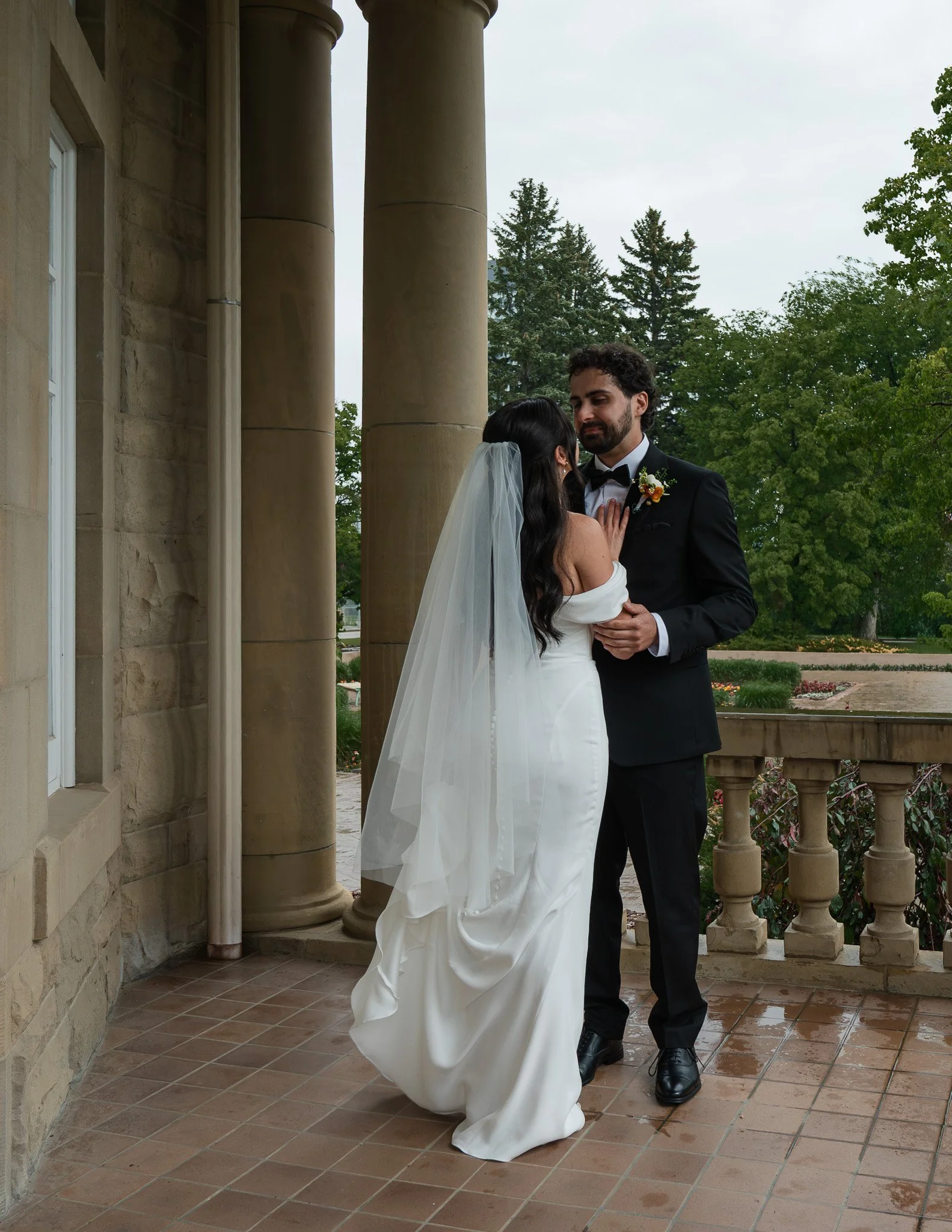 A bride and groom sharing an intimate moment outside a stone building with columns, surrounded by trees and greenery. The bride is wearing a white wedding dress with a veil, and the groom is dressed in a black tuxedo with a boutonniere.