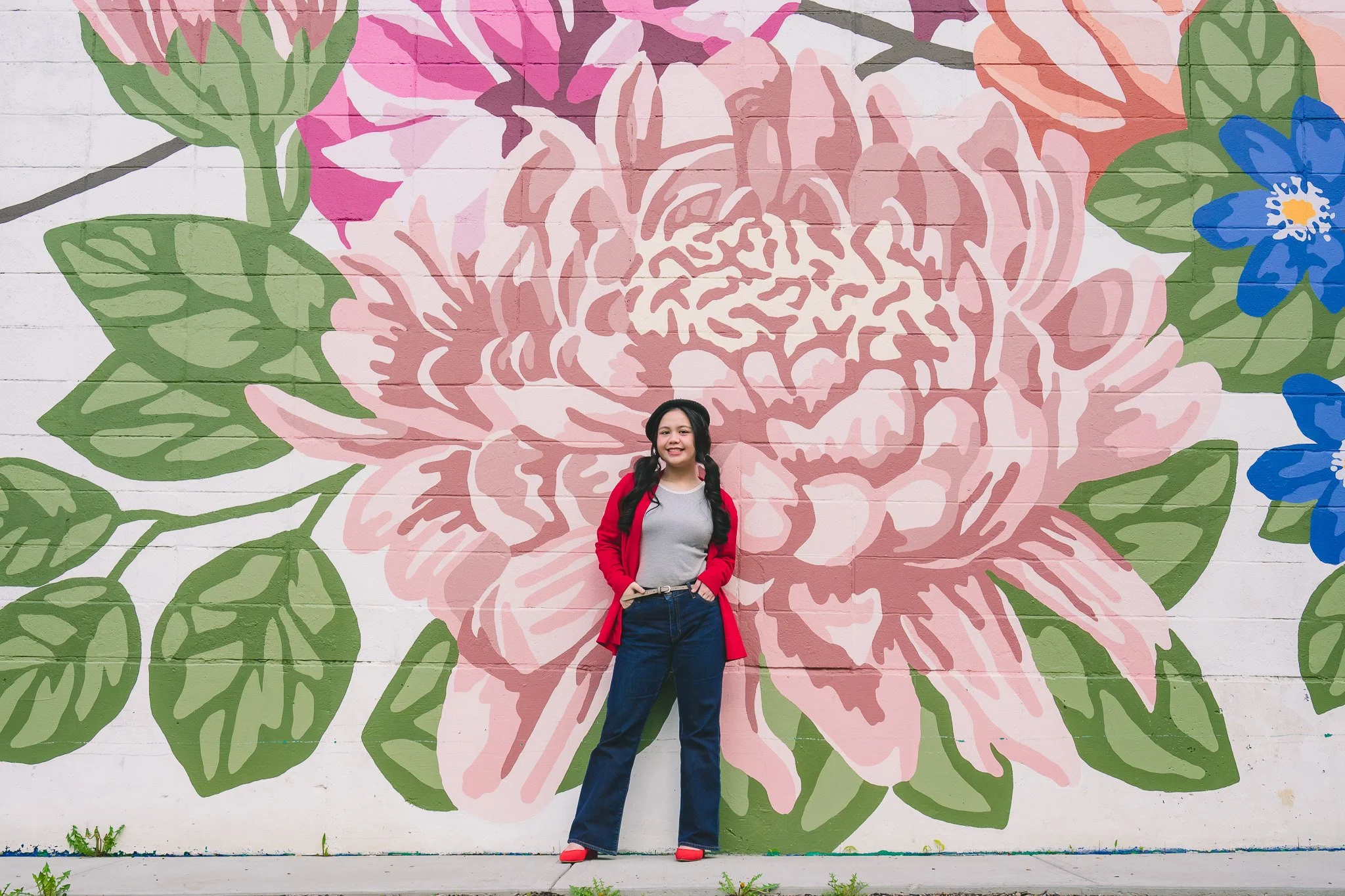 A young girl with long dark hair, wearing a gray shirt, blue jeans, red jacket, and red shoes, standing in front of a large floral mural painted on a brick wall. The mural features a pink flower with green leaves and smaller blue flowers.